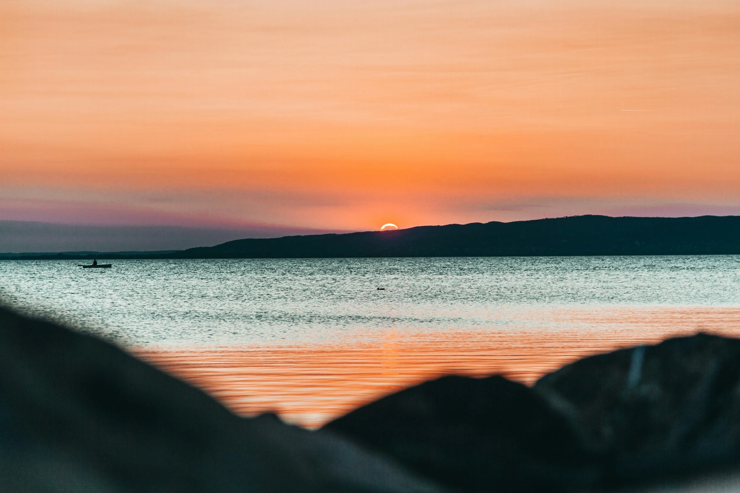 Golden and pink sunset over the calm waters of Lake Balaton in Hungary, viewed from a stone shoreline with low hills silhouetted on the far bank
