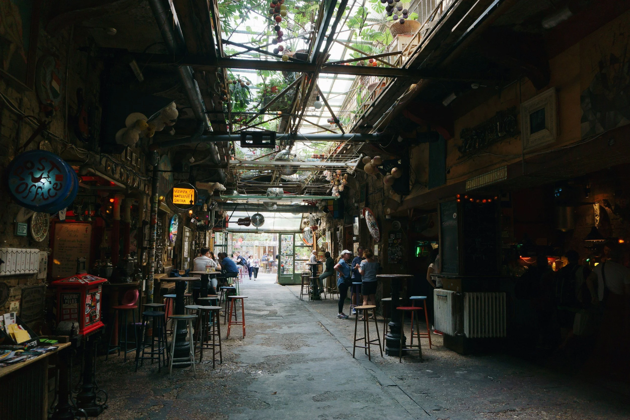 Dimly lit interior courtyard of a Budapest ruin bar with mismatched furniture, industrial pipes, hanging plants, and guests seated at bar tables beneath a glass roof