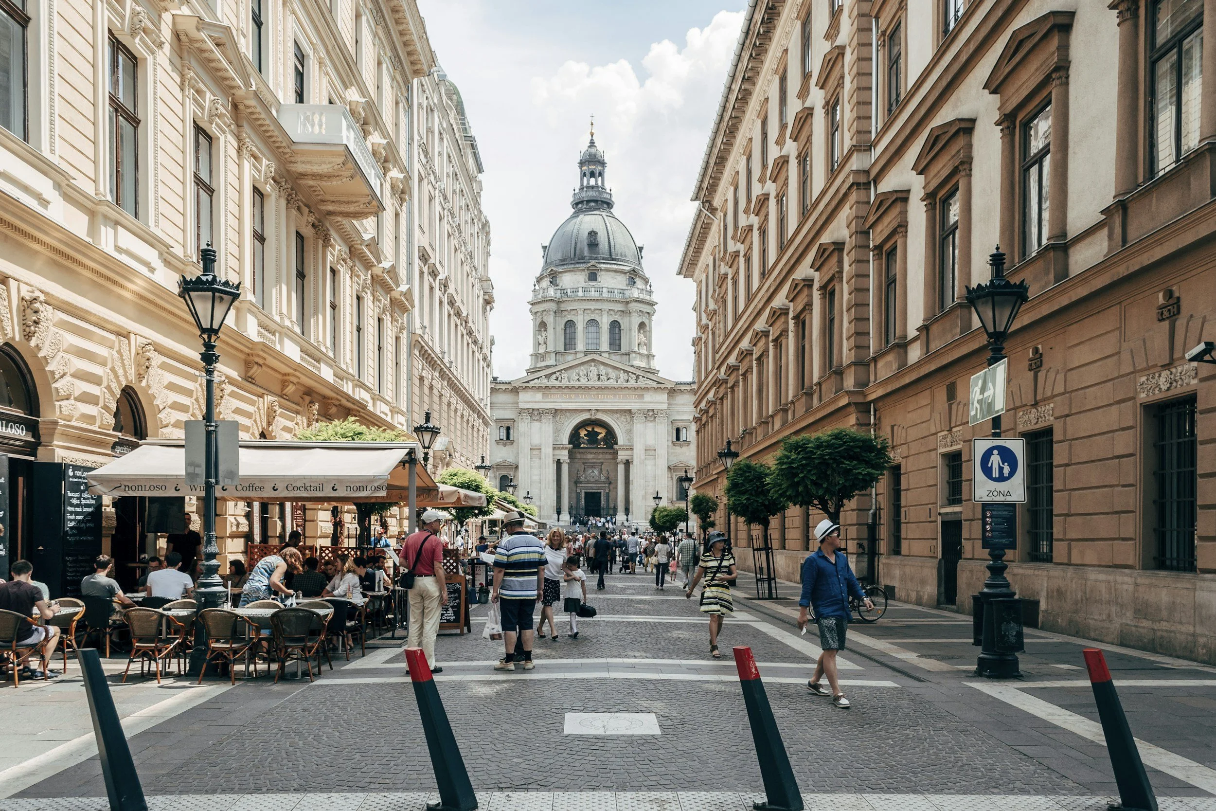 Tree-lined boulevard in Pest with grand 19th-century buildings, street cafes, and the dome of St Stephen's Basilica visible at the end of the street in Budapest, Hungary