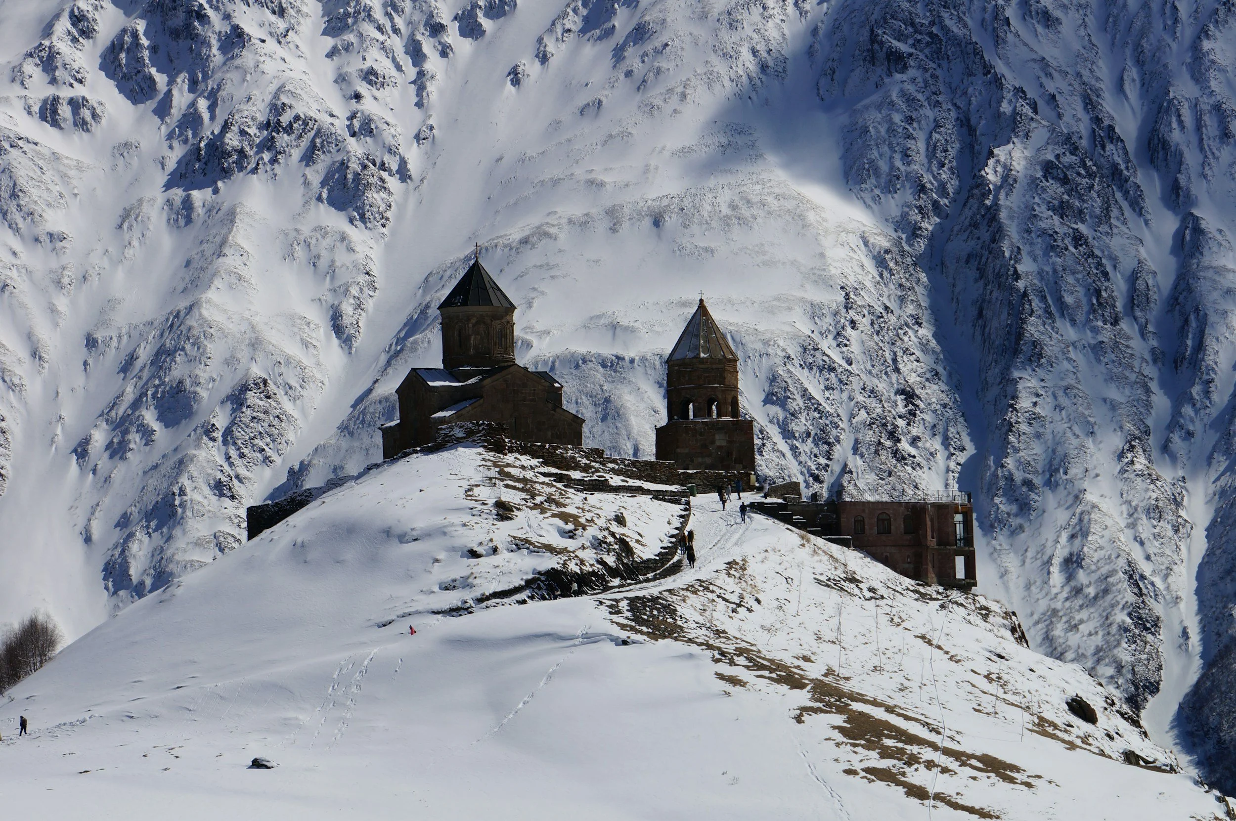 Gergeti Trinity Church and its bell tower standing in deep snow on a steep ridge, surrounded by sheer snow-covered Caucasus mountain walls near Kazbegi in winter, Georgia