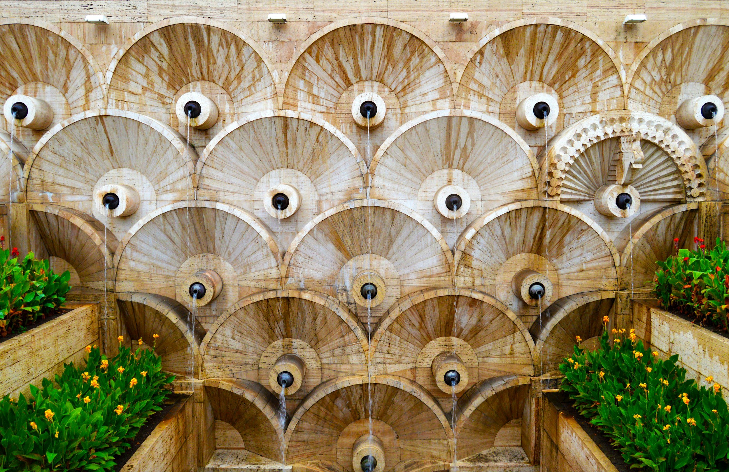 Close-up of layered fan-shaped stone fountain tiers with black sphere spouts at the Cascade complex in Yerevan, flanked by orange and red flowering plants in summer, Armenia