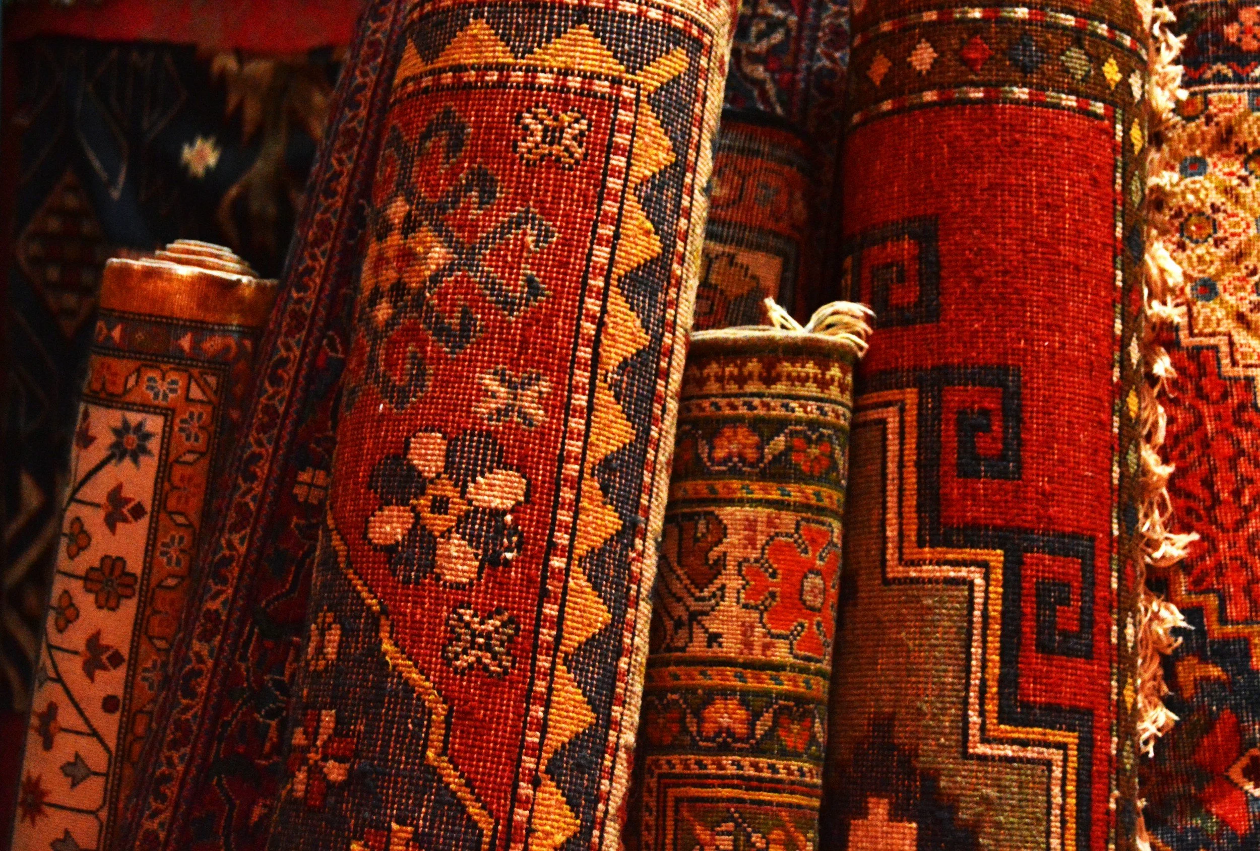 Close-up of rolled traditional Armenian handwoven carpets stacked together, showing rich red tones, intricate floral and geometric motifs and gold border patterns in warm low light