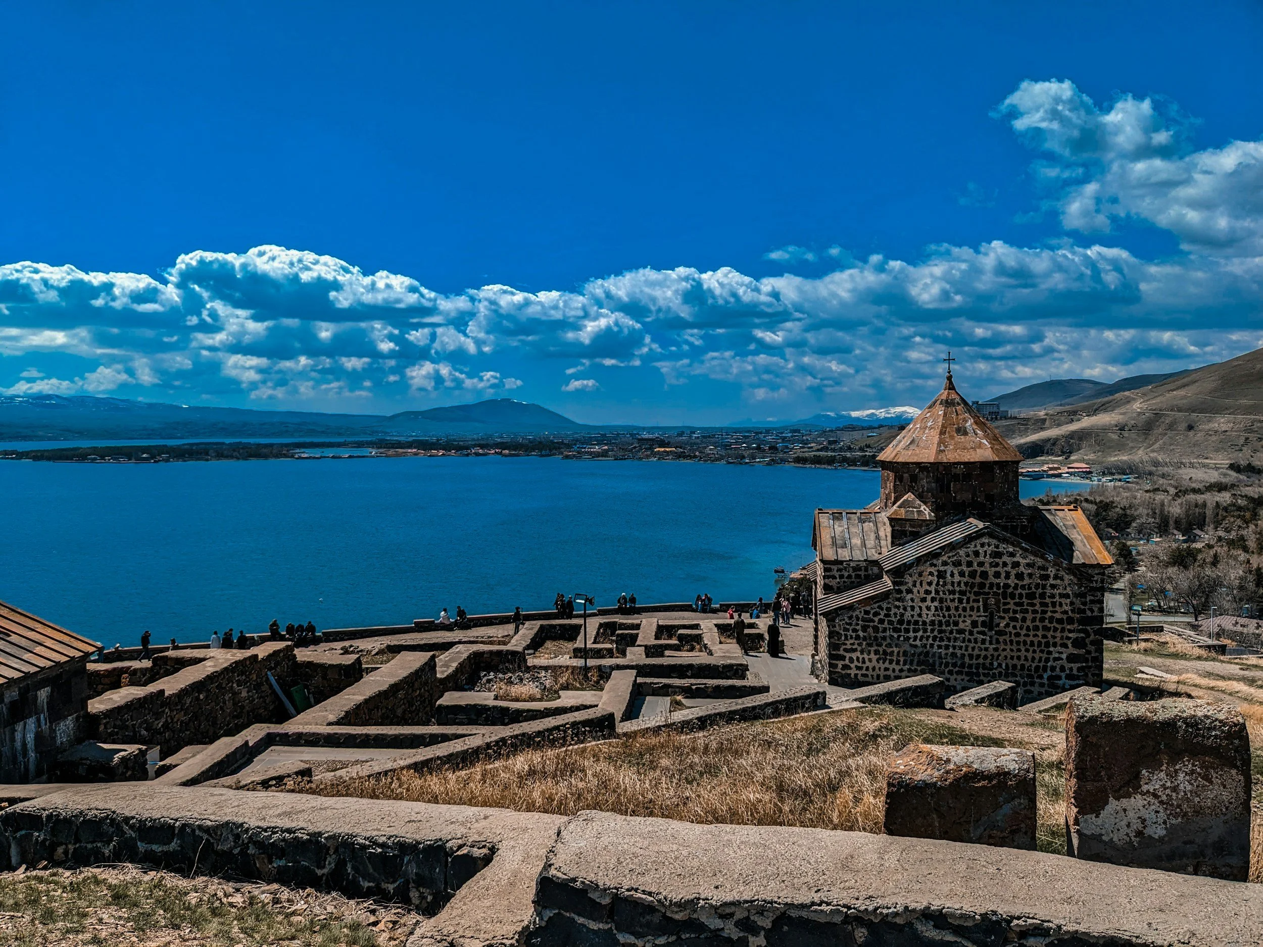 Sevanavank monastery church with its copper-toned roof on a rocky promontory above the deep blue waters of Lake Sevan, with ancient stone wall ruins in the foreground and snow-capped mountains on the far shore, Armenia