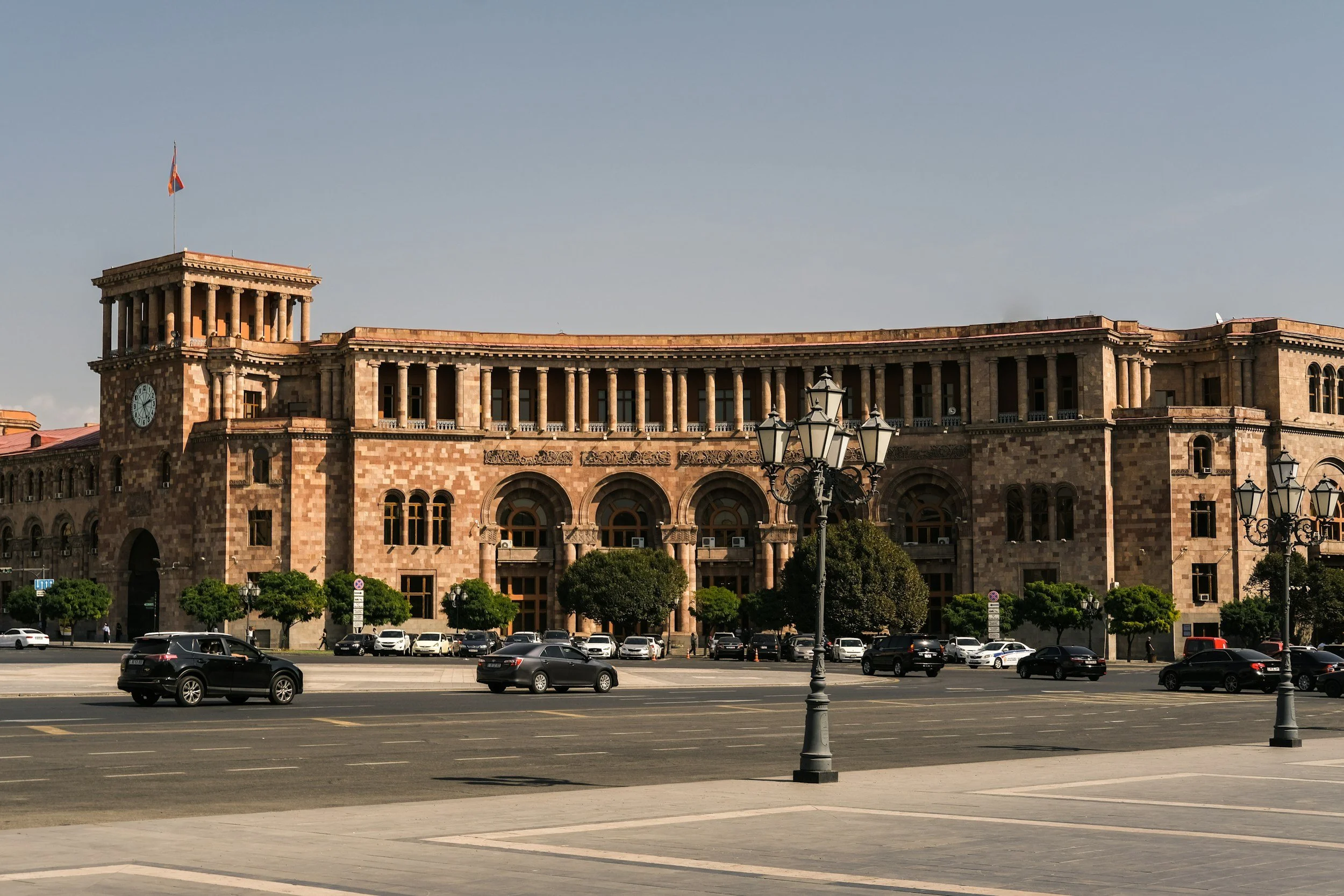 Grand neoclassical government building built from pink and golden tuff stone facing Republic Square in central Yerevan, with colonnaded arcades, a clock tower, ornamental carvings and an Armenian flag flying above