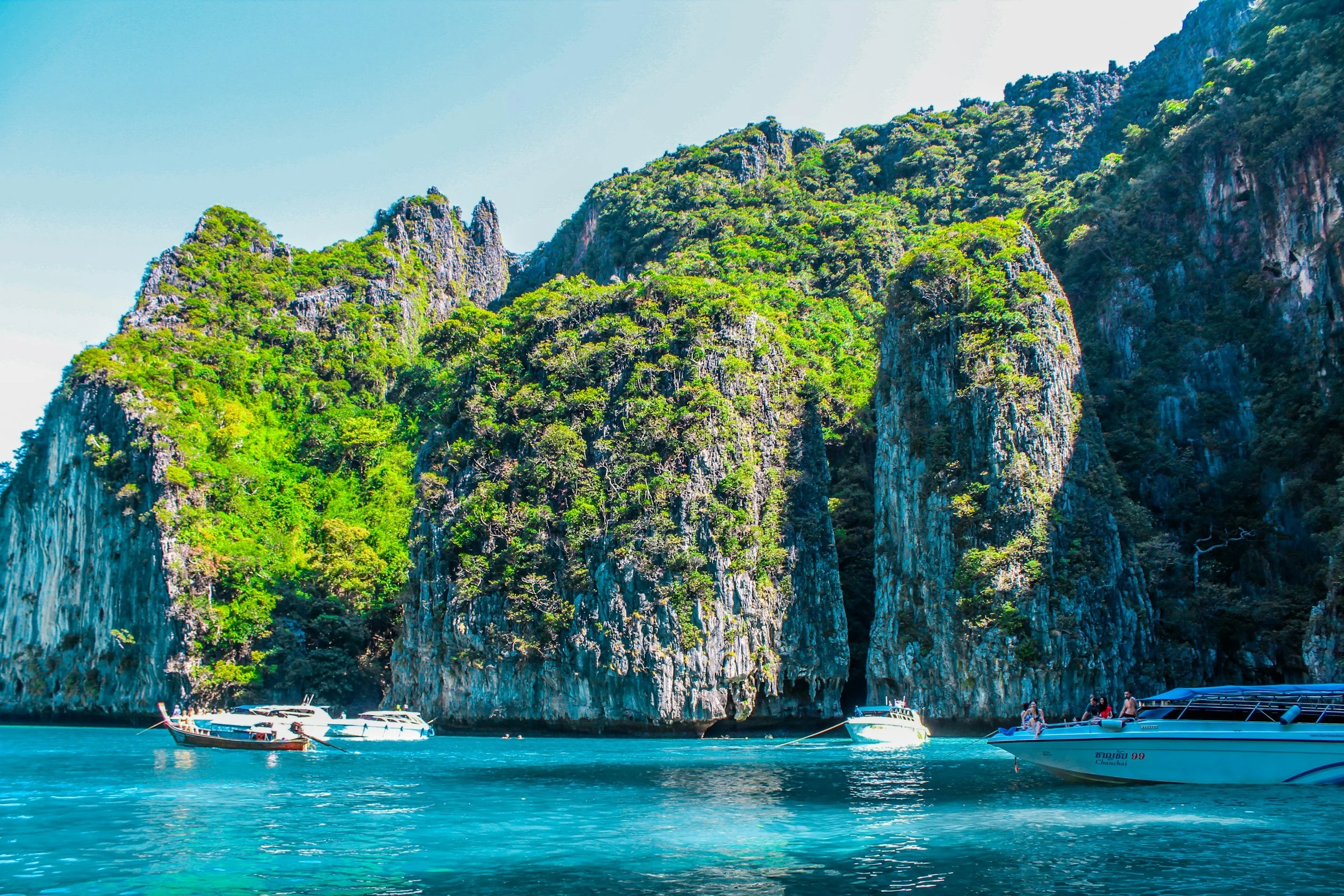 Towering jungle-covered limestone cliffs rising from turquoise water near Phuket in the Andaman Sea, Thailand, with longtail boats moored at the base of the cliffs