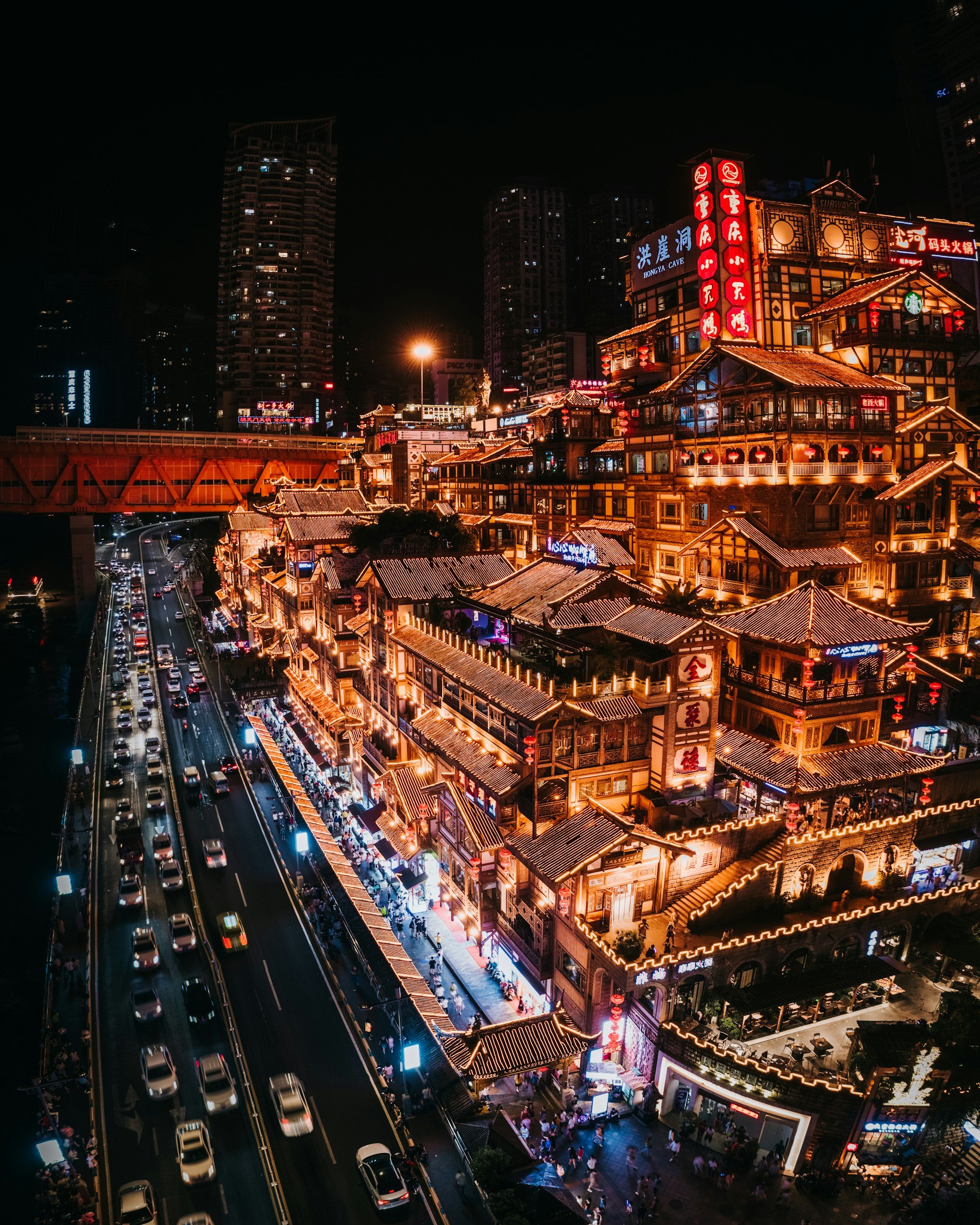 Aerial night view of the Hongyadong stilt house district in Chongqing, China, with its stacked traditional wooden buildings illuminated by warm neon lights cascading down the cliff face above a busy road