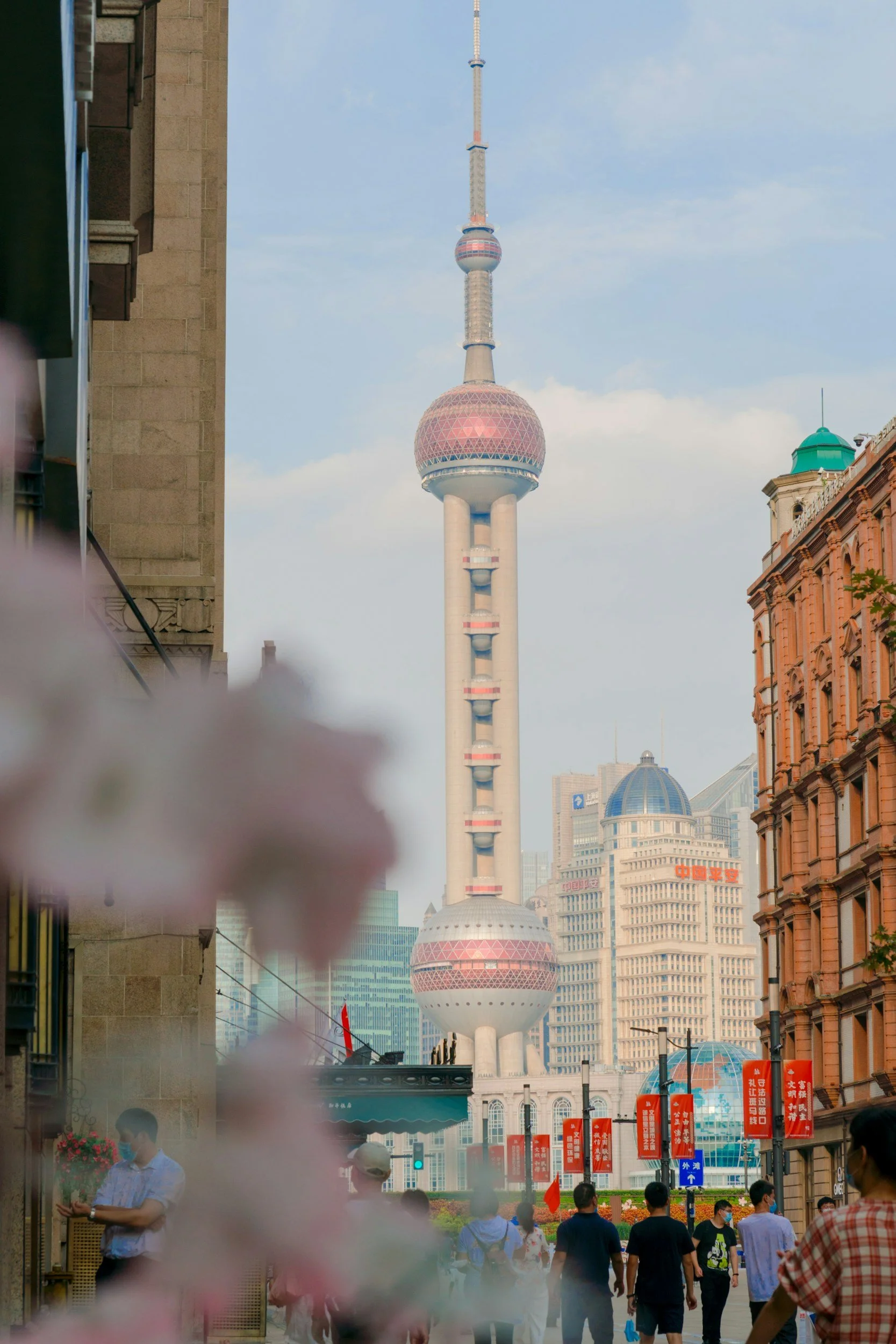 The Oriental Pearl Tower visible through cherry blossom alongside ornate European Art Deco and neoclassical buildings on the Shanghai Bund, reflecting the city's layered immigration and colonial history, China