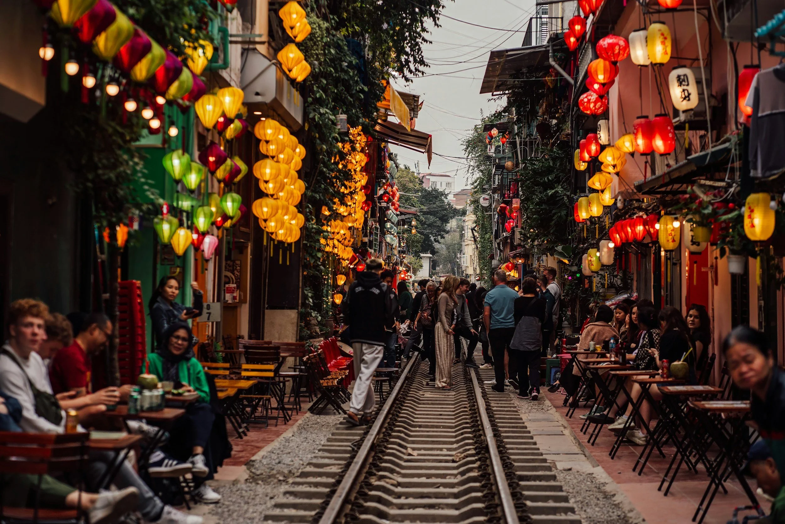 The famous Train Street in Hanoi's Old Quarter lined with colourful silk lanterns, outdoor cafe tables and shopfronts on both sides, with visitors gathered along the narrow railway track at dusk