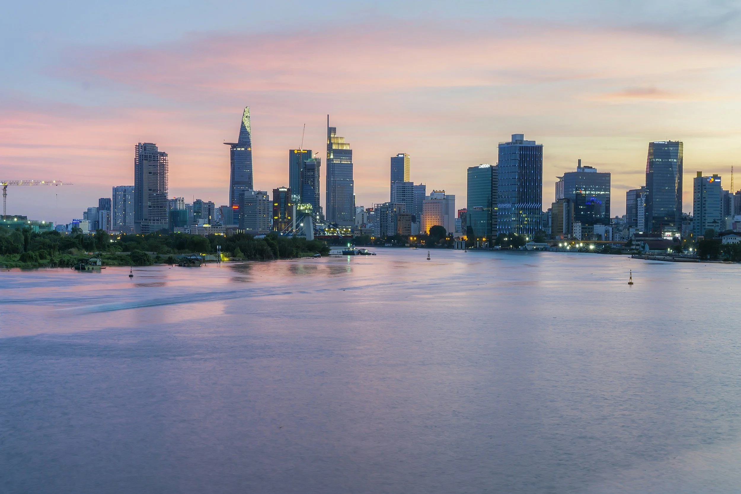The Ho Chi Minh City skyline at dusk showing the Bitexco Financial Tower and modern high-rises reflected in the calm Saigon River under a pink and purple sky, Vietnam