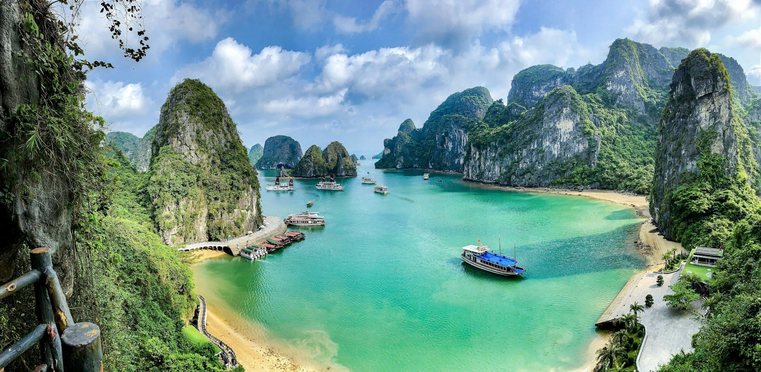 A private wooden junk cruise boat anchored in turquoise water surrounded by towering limestone karst islands in Ha Long Bay, Vietnam, with a pier and smaller boats visible in the foreground