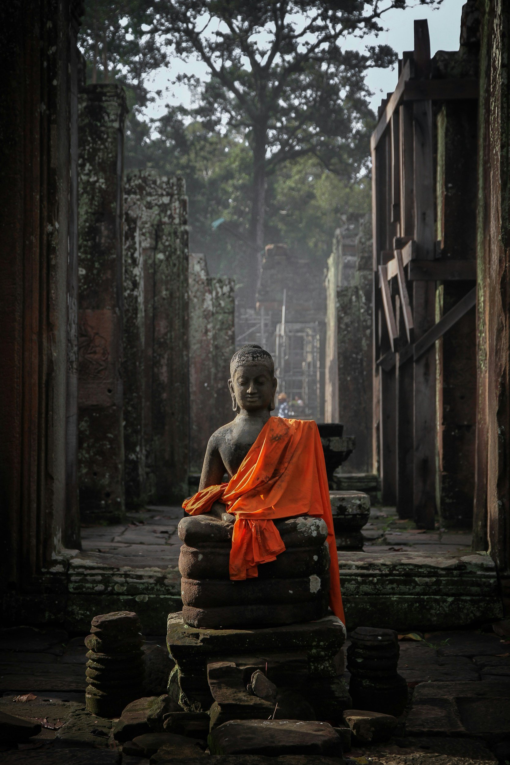 Ancient moss-covered stone Buddha statue draped in an orange saffron robe inside a dimly lit sandstone temple chamber at the Angkor archaeological complex, Siem Reap, Cambodia