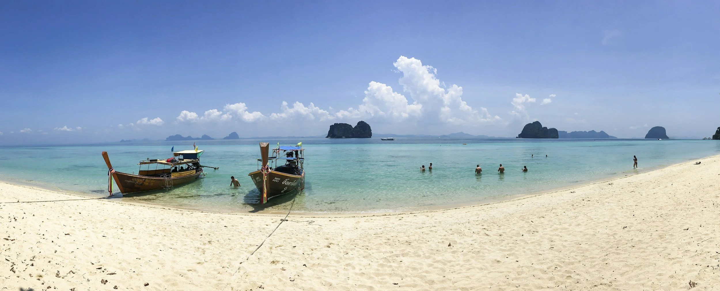 A wooden longtail boat moored on a white sand beach with clear turquoise water, swimmers visible in the distance and a limestone island rising on the horizon in the Gulf of Thailand, Cambodia
