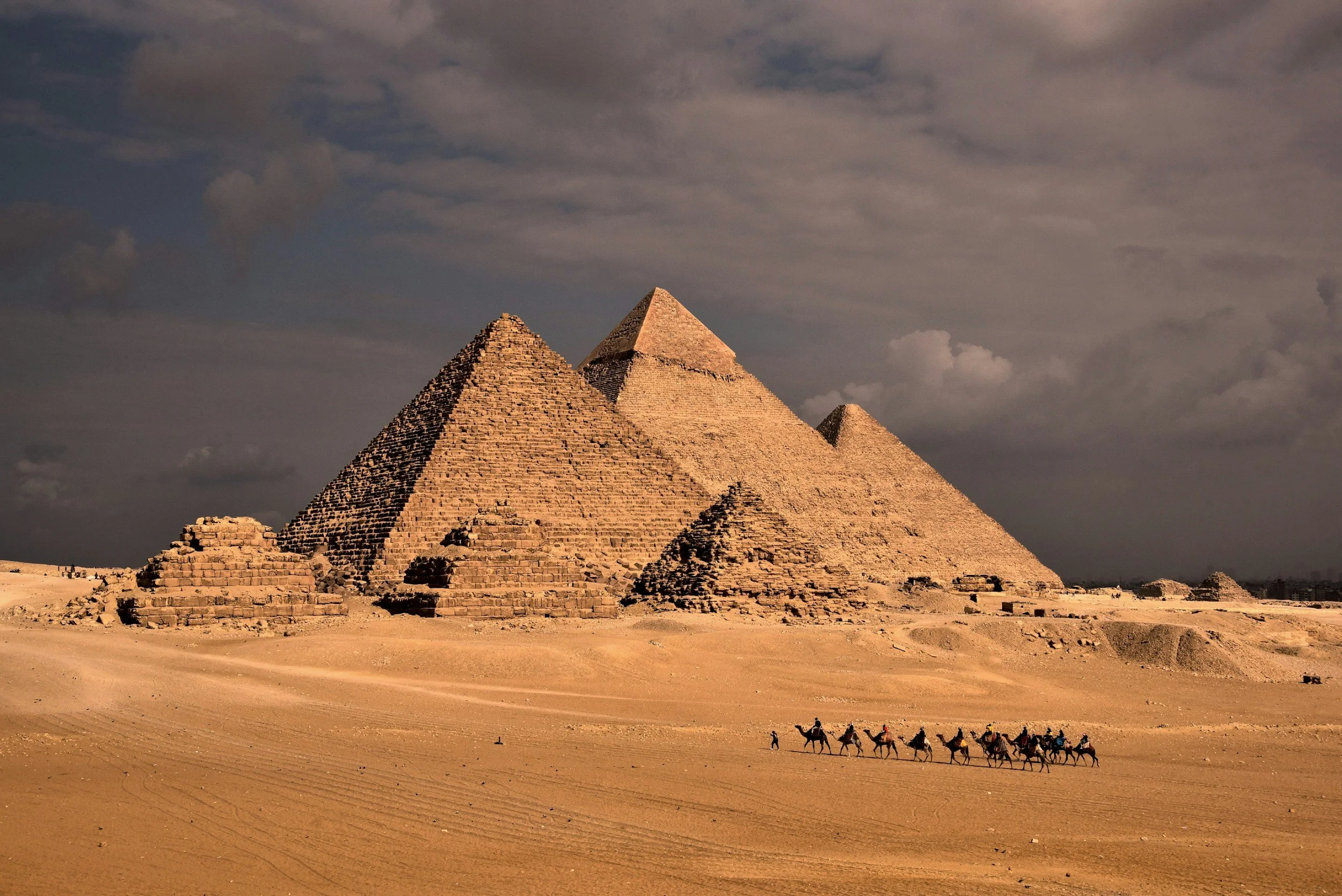 The three Pyramids of Giza rising above the desert under a dramatic sky with camels and riders in the foreground, Egypt