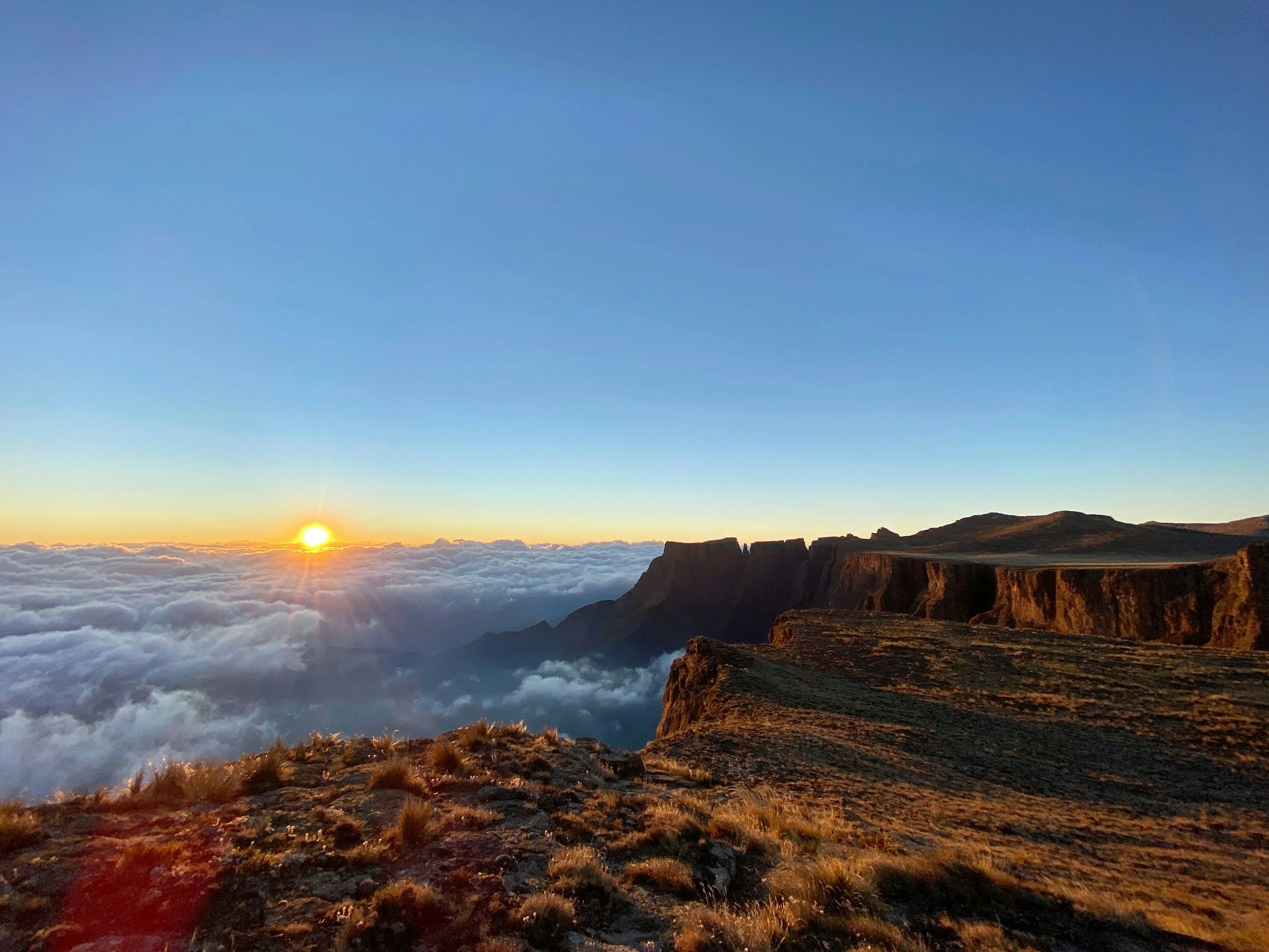 Sun rising over dramatic rocky cliffs in the Drakensberg mountains, with a sea of clouds below and a vivid orange sky, KwaZulu-Natal, South Africa