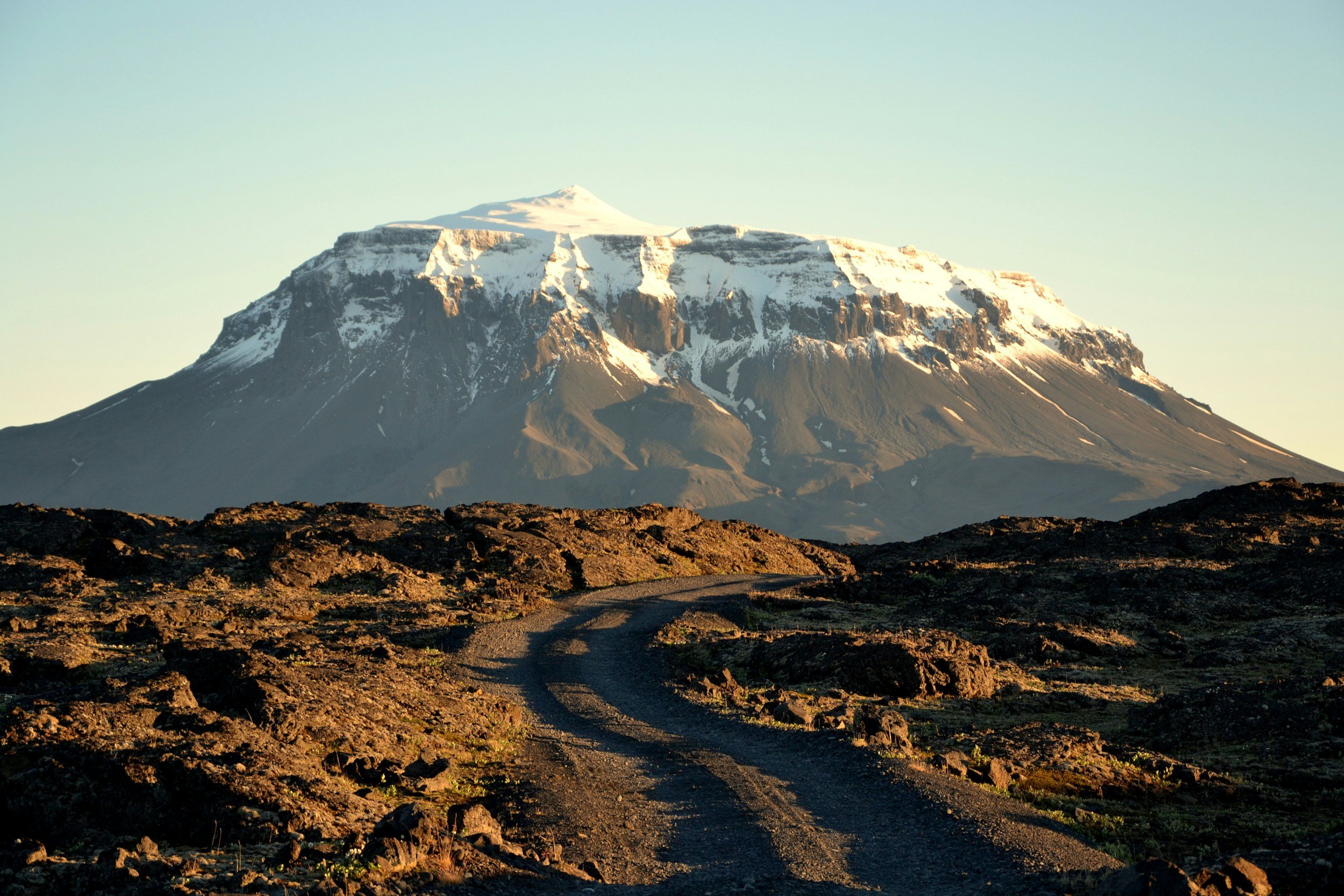 Winding dirt road through volcanic lava fields with the snow-capped summit of Mount Kilimanjaro rising above the clouds in the background, Amboseli, Kenya