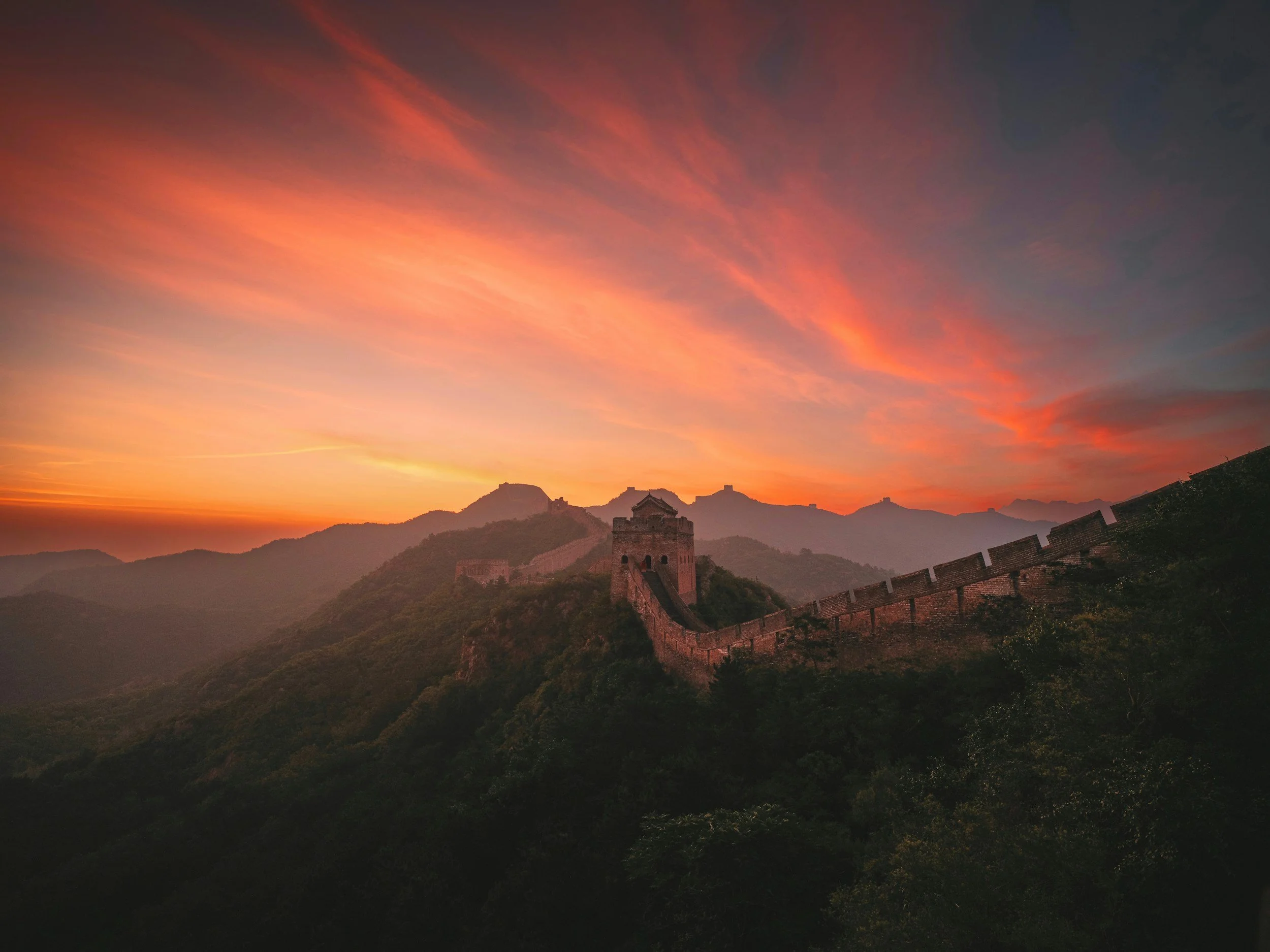 The Great Wall of China silhouetted against a dramatic red and orange sunset sky, with the wall curving across forested mountain ridges and a lone watchtower at its centre