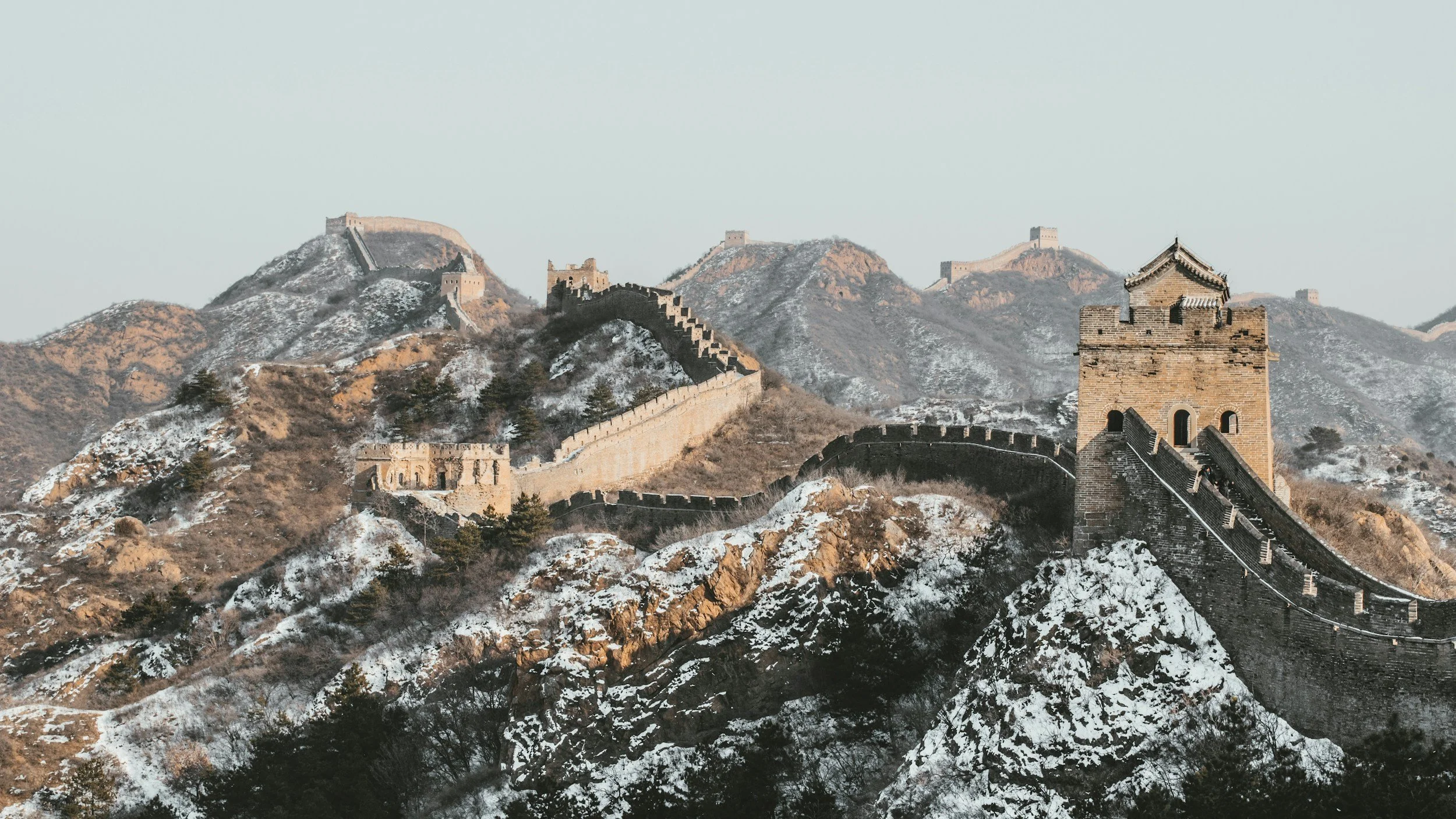 Wide panoramic view of the Great Wall of China in winter, with snow on the mountains, a restored watchtower rising to the right and a ruined section visible in the middle distance