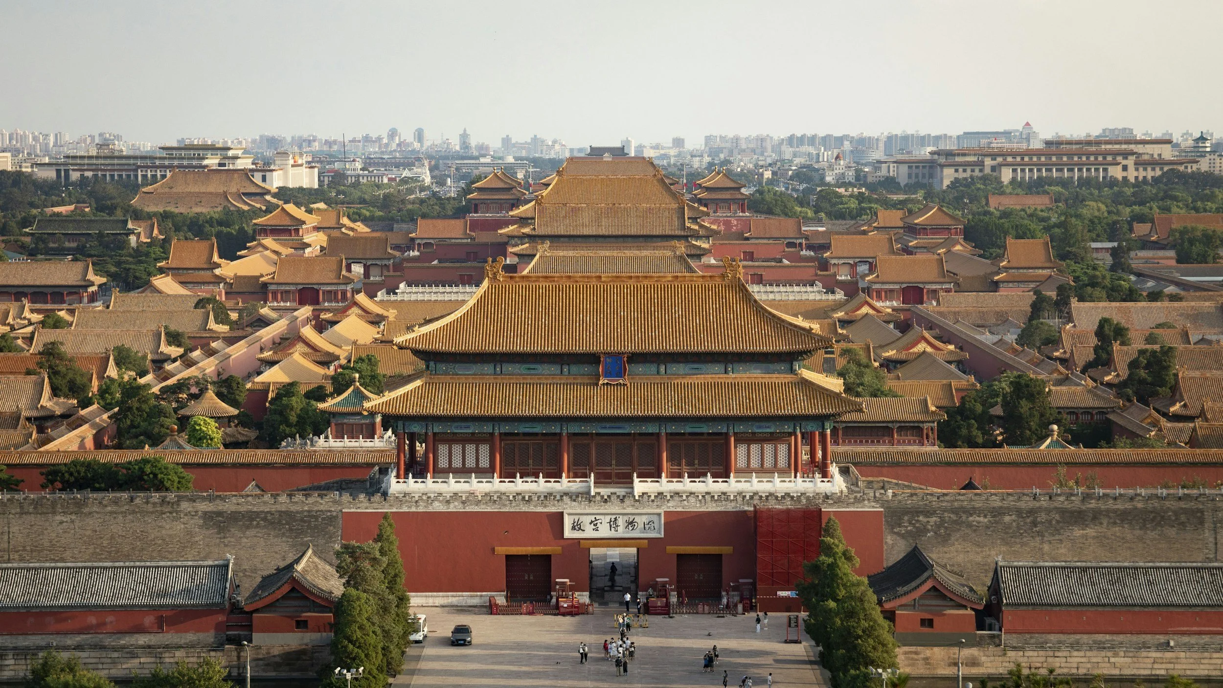 Aerial view of the Forbidden City in Beijing, showing the vast complex of red walls and golden-tiled imperial rooftops with the modern city skyline visible beyond