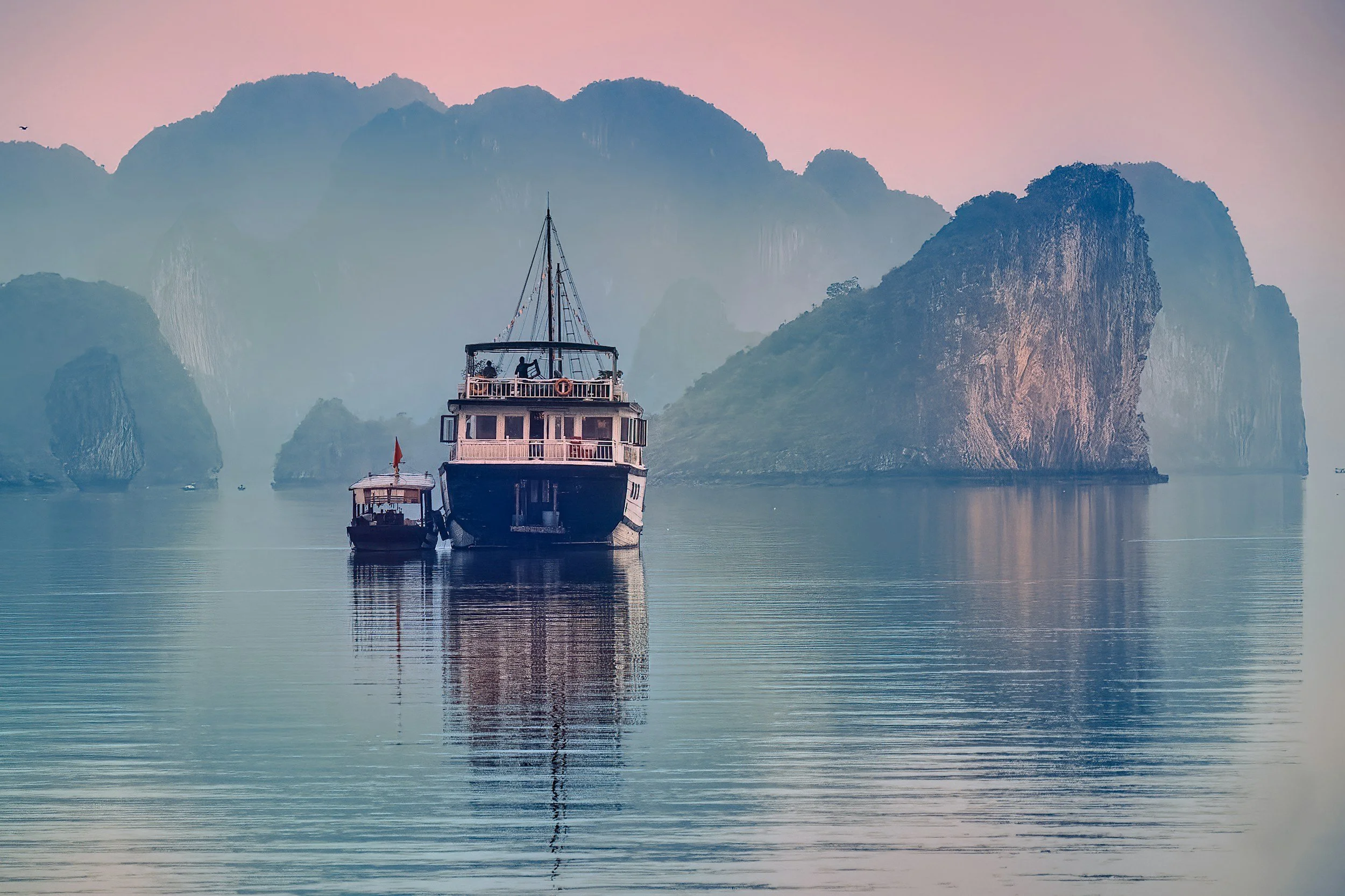 A private junk boat floating in the still waters of Ha Long Bay at dawn, with limestone karsts emerging from the morning mist under a soft pink sky, Vietnam