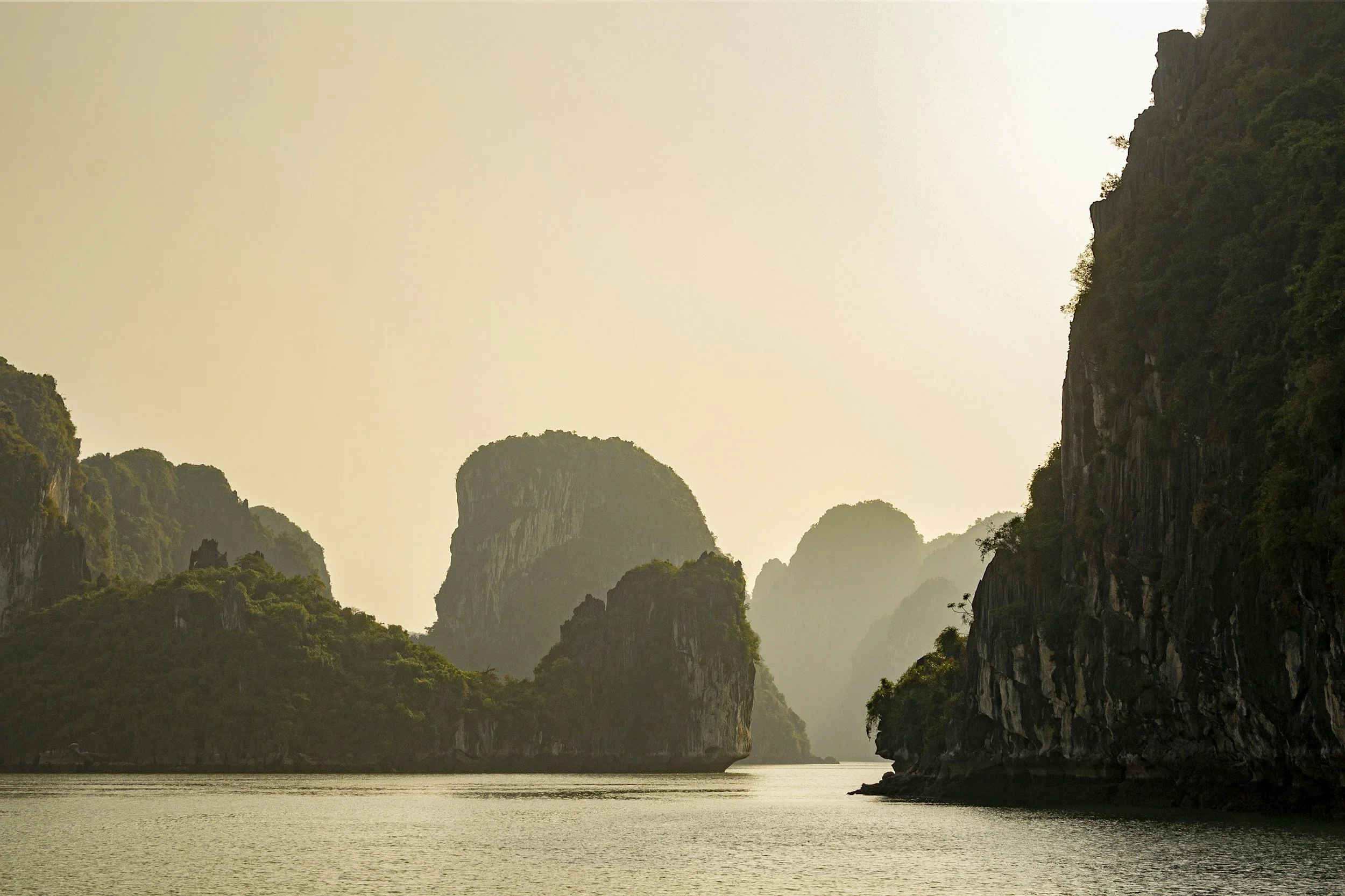 Dramatic limestone karsts rising from the glassy still waters of Ha Long Bay in warm hazy afternoon light, Vietnam