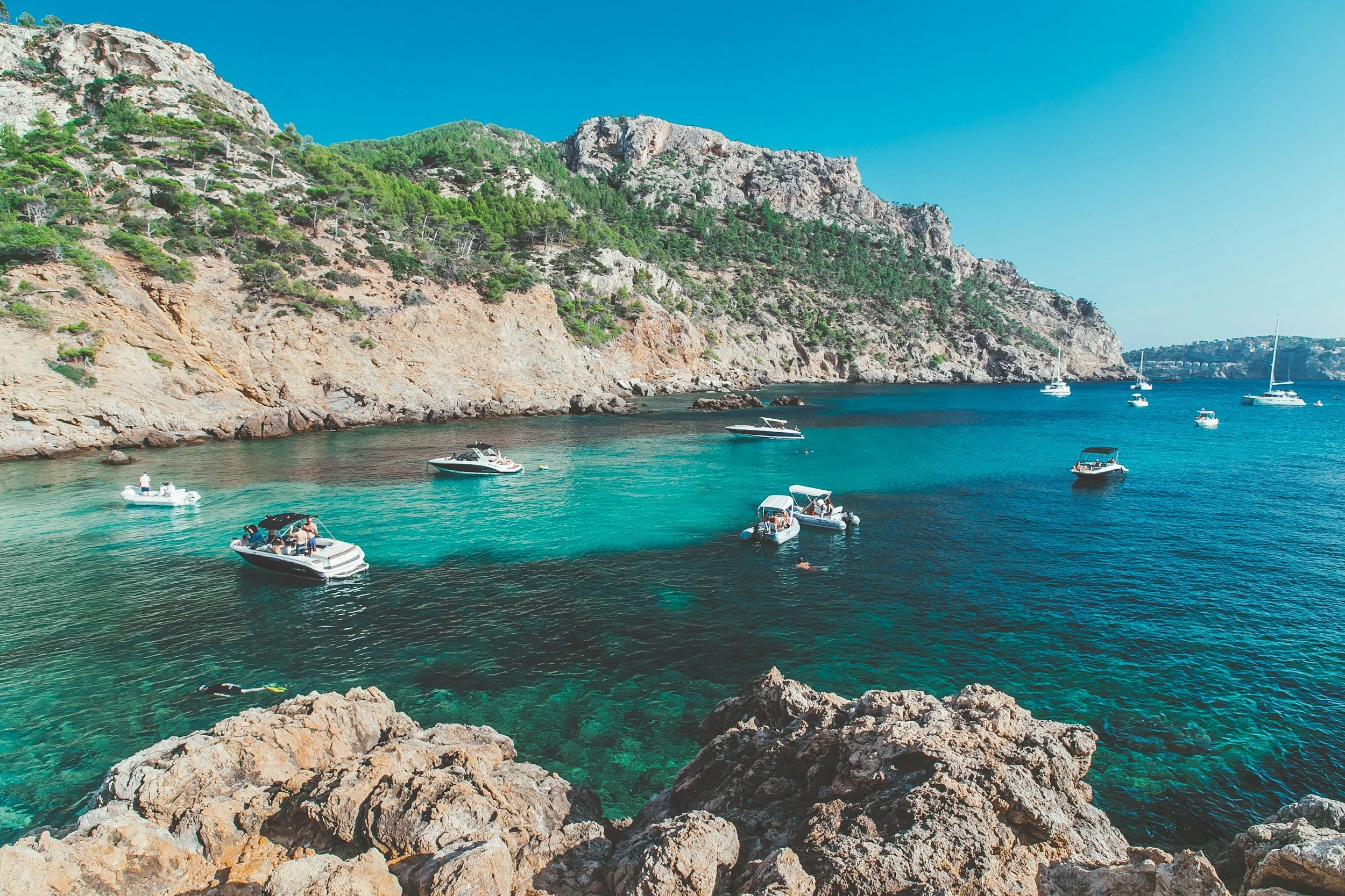 Turquoise waters in a rocky cove in the Balearic Islands with sailing boats at anchor and pine-covered limestone cliffs rising on either side, Spain