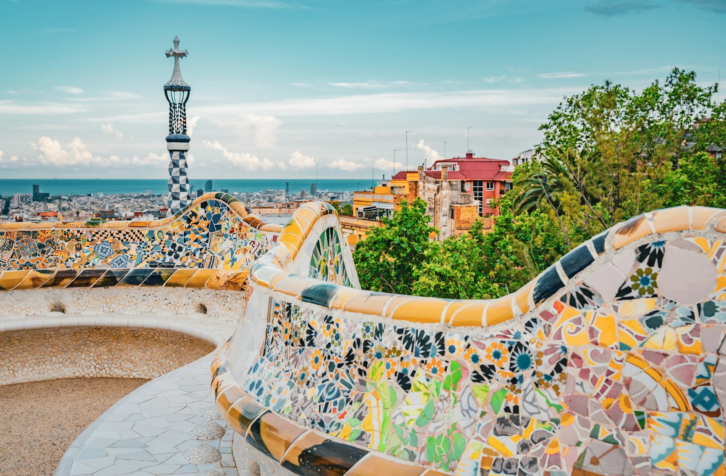 The undulating mosaic-tiled bench terrace of Park Güell by Antoni Gaudí, with the checkered tower and views over Barcelona's rooftops and the Mediterranean Sea beyond, Spain