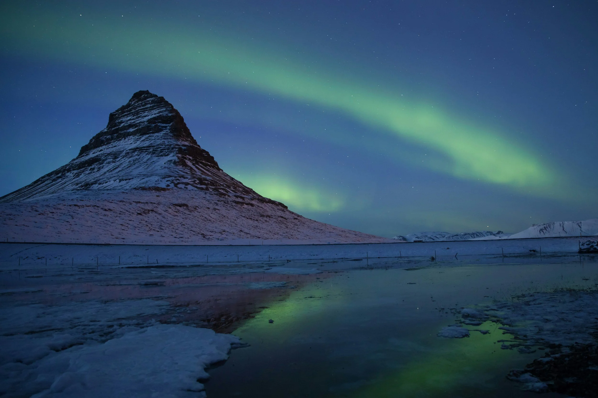 Green aurora borealis streaming across the night sky above the snow-covered cone of Kirkjufell mountain in the Snæfellsnes Peninsula, reflected in still water below, Iceland