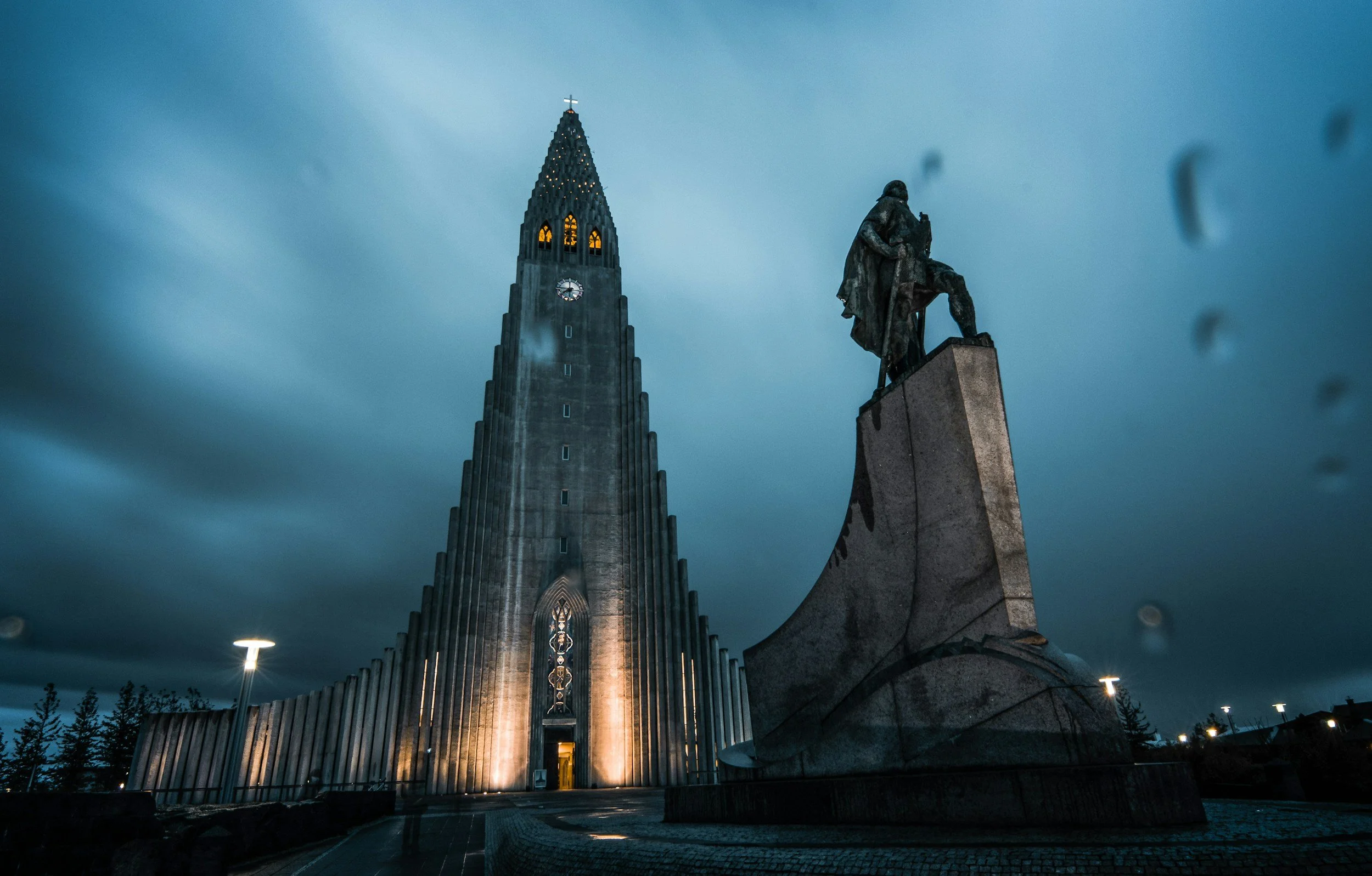 Hallgrímskirkja church illuminated at night in Reykjavík, with the statue of Leif Eriksson in the foreground and dramatic stormy clouds moving across a dark blue sky, Iceland
