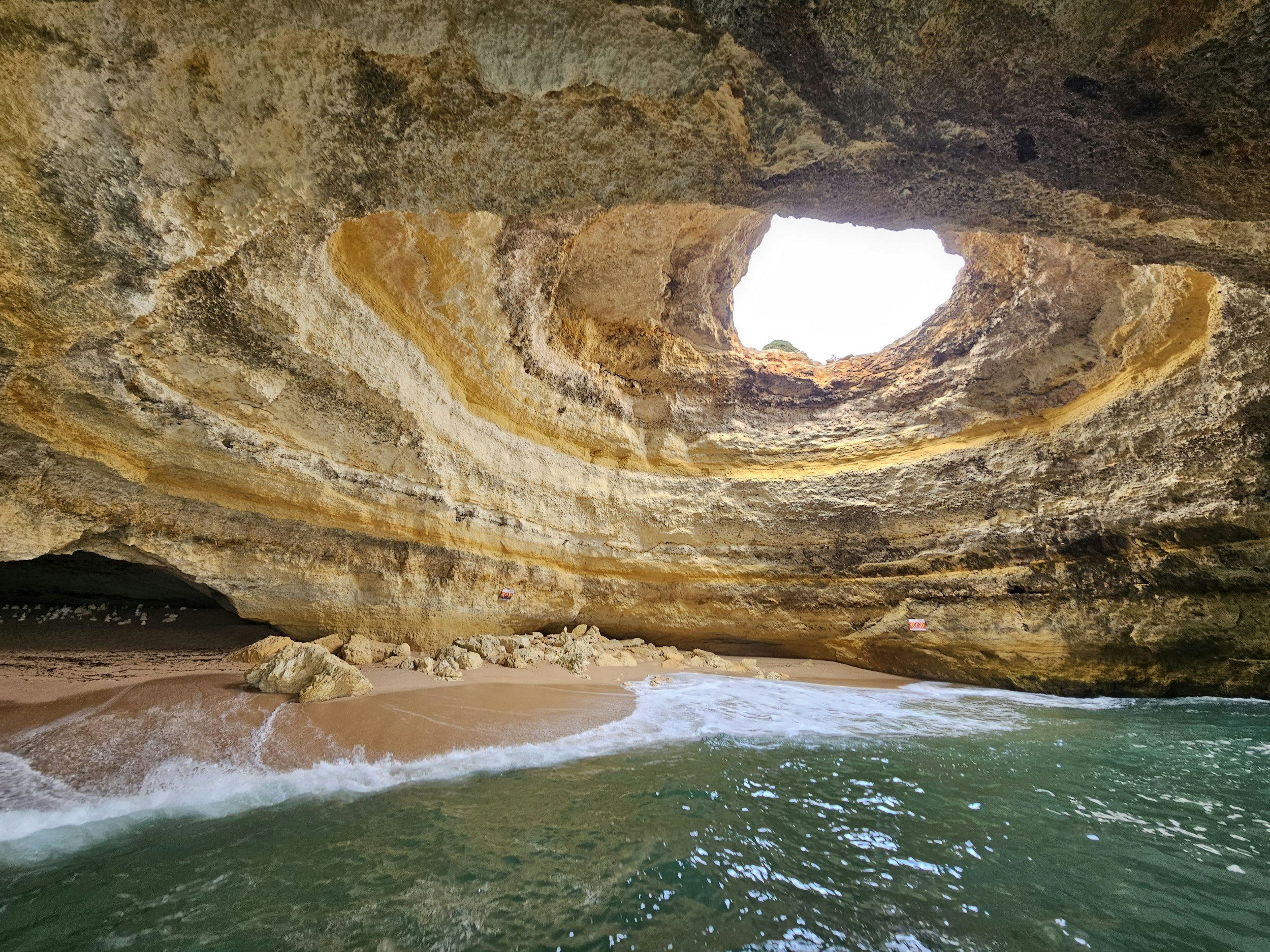 Inside the famous Benagil sea cave in the Algarve, looking up through the natural domed ceiling opening to the sky, with waves washing onto the sandy beach below, Portugal