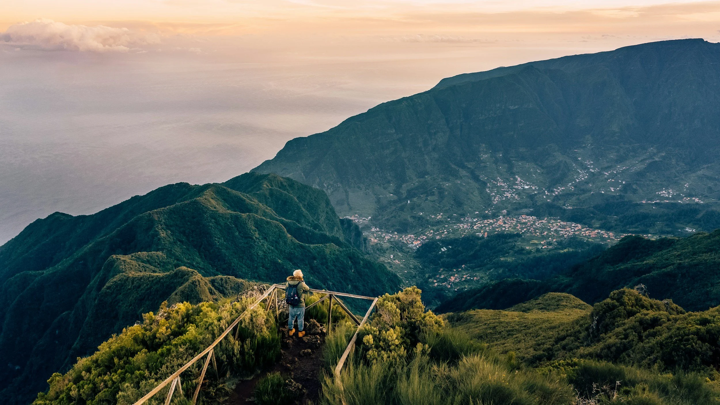 A hiker walking along a narrow levada irrigation channel trail through lush green mountain scenery in Madeira, with dramatic valley views and the Atlantic beyond, Portugal