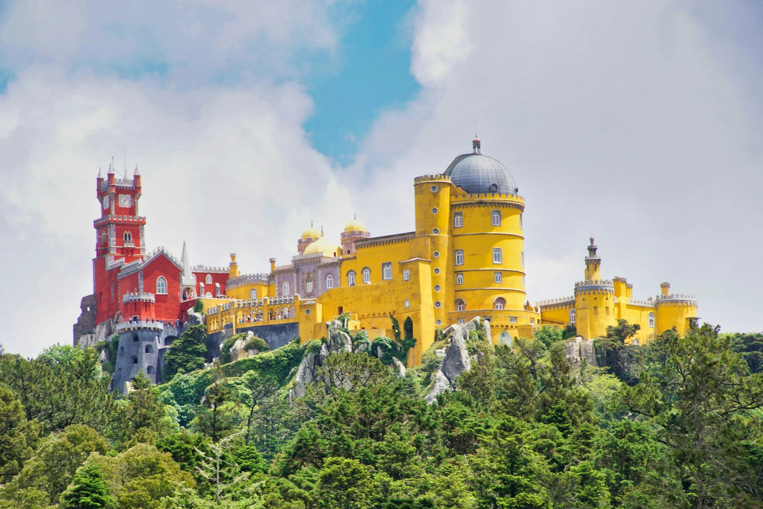 The colourful romantic Pena Palace perched on a forested hilltop in Sintra, with its yellow and red towers rising above the trees under a blue sky, Portugal