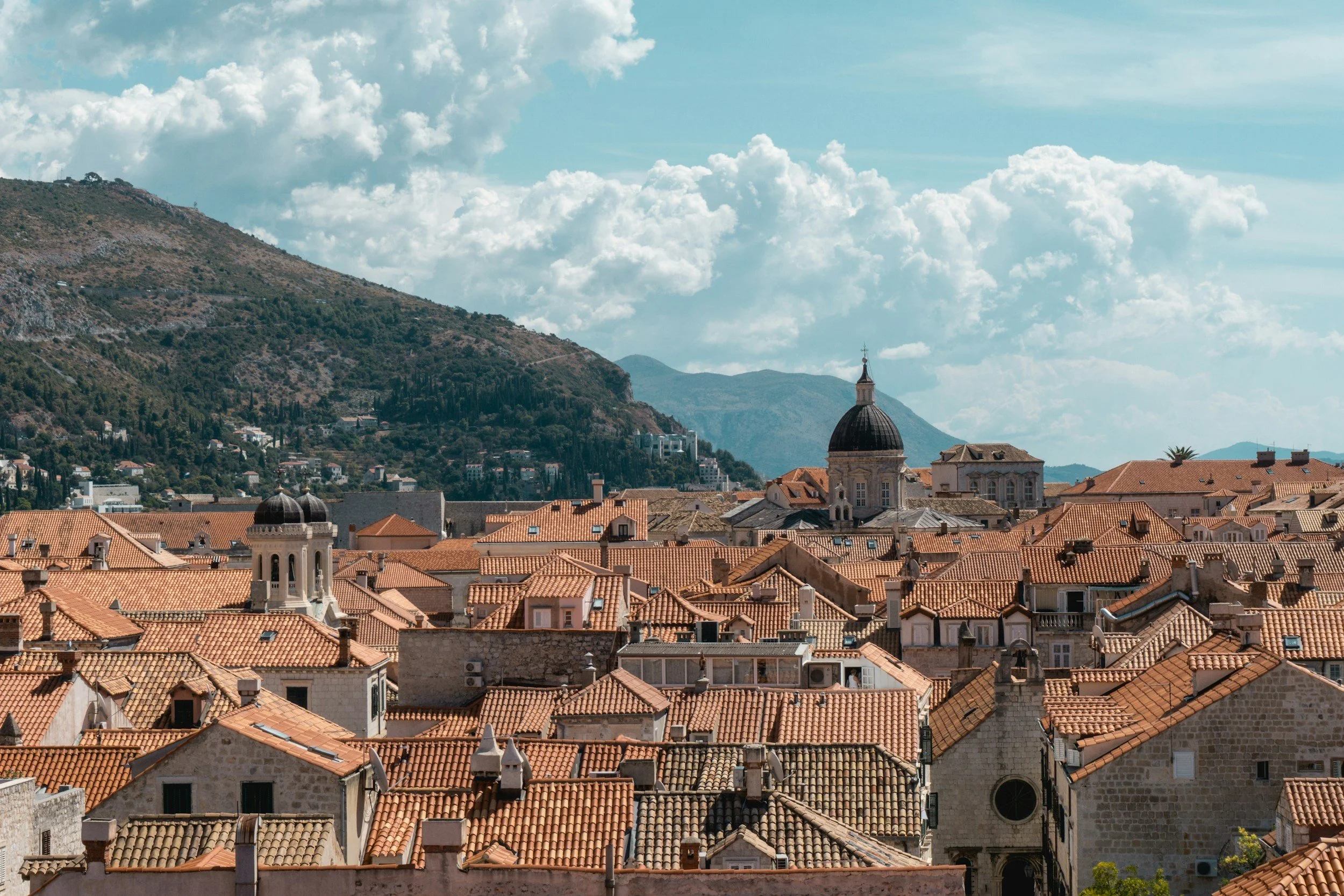Terracotta rooftops of Dubrovnik's medieval old town with the baroque cathedral dome, surrounding mountains and the blue Adriatic Sea visible in the background, Croatia