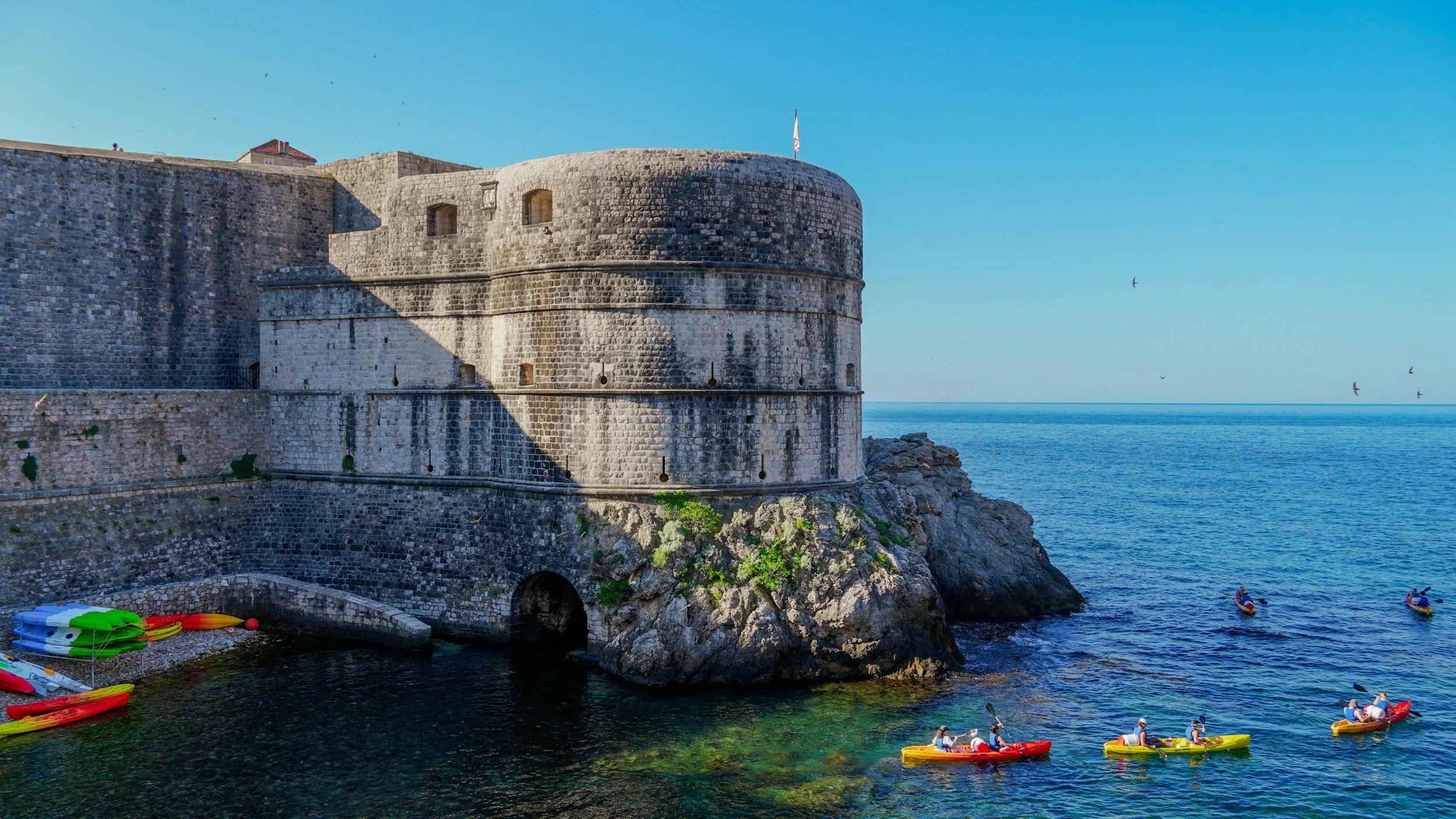 Sea kayakers paddling past the base of Dubrovnik's ancient limestone city walls on the clear blue Adriatic, with the medieval Bokar Tower rising above them, Croatia