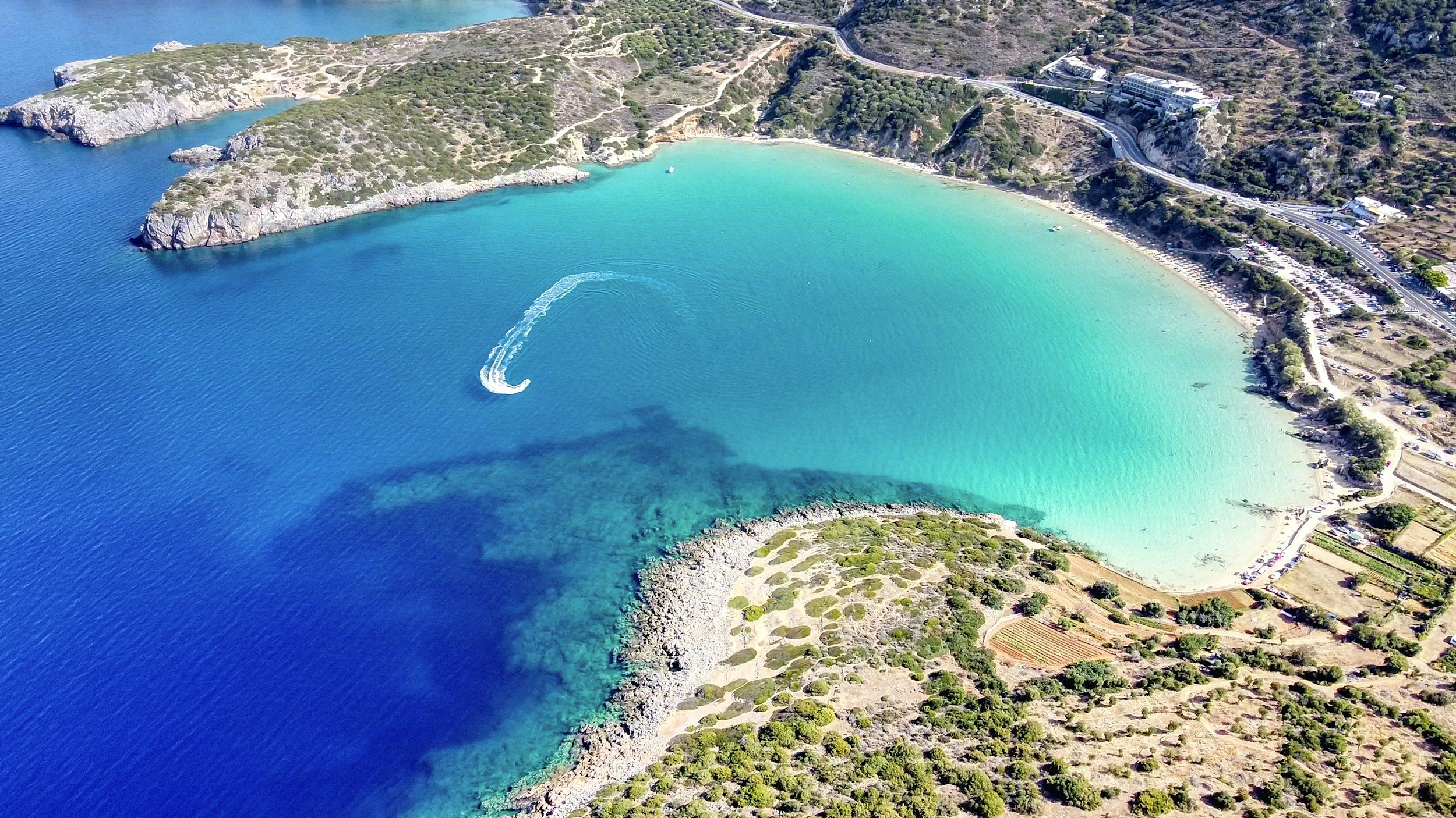 Aerial view of a deep turquoise bay with rocky coastline and a speedboat leaving a white wake on the water, on the island of Crete, Greece