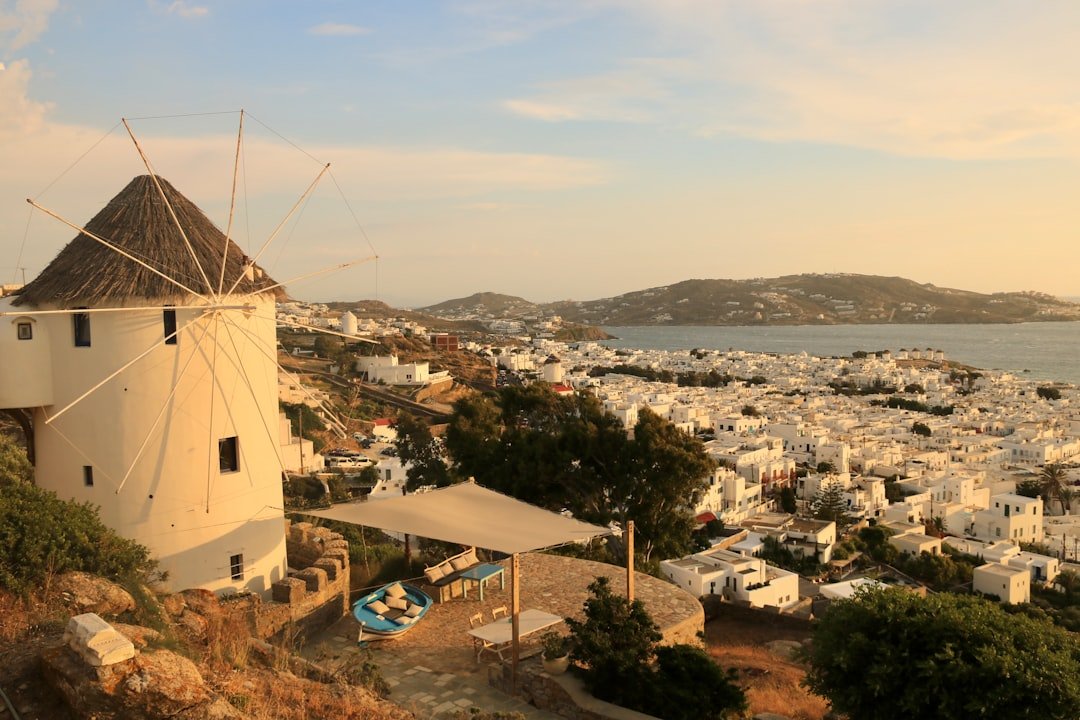 A traditional white windmill overlooking the town and harbour of Mykonos at sunset, with the Aegean Sea and distant hills in the background, Greece