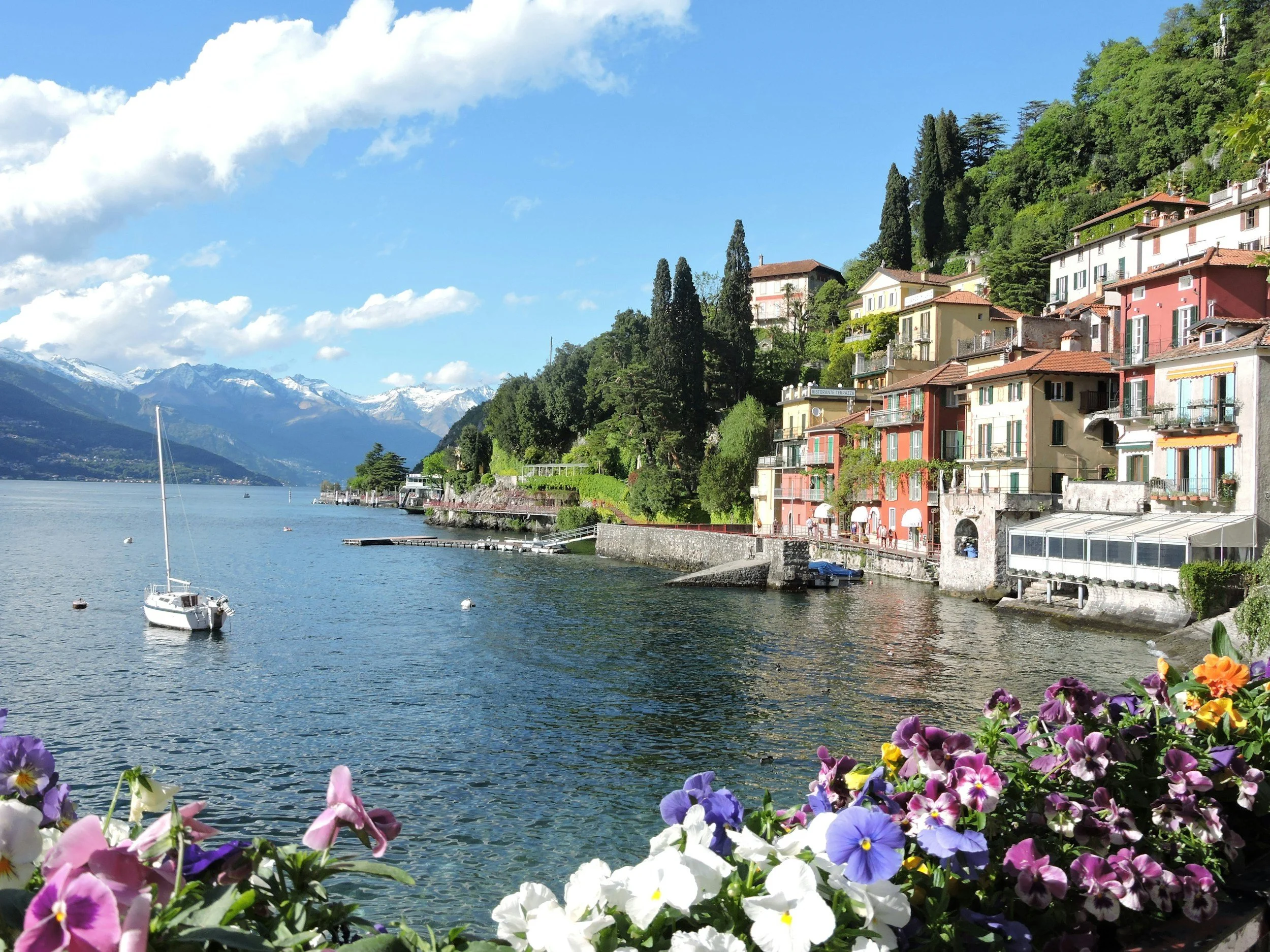 The lakeside promenade at Varenna on Lake Como with colourful flowers, historic villas and snow-capped Alps rising beyond the deep blue water, Italy