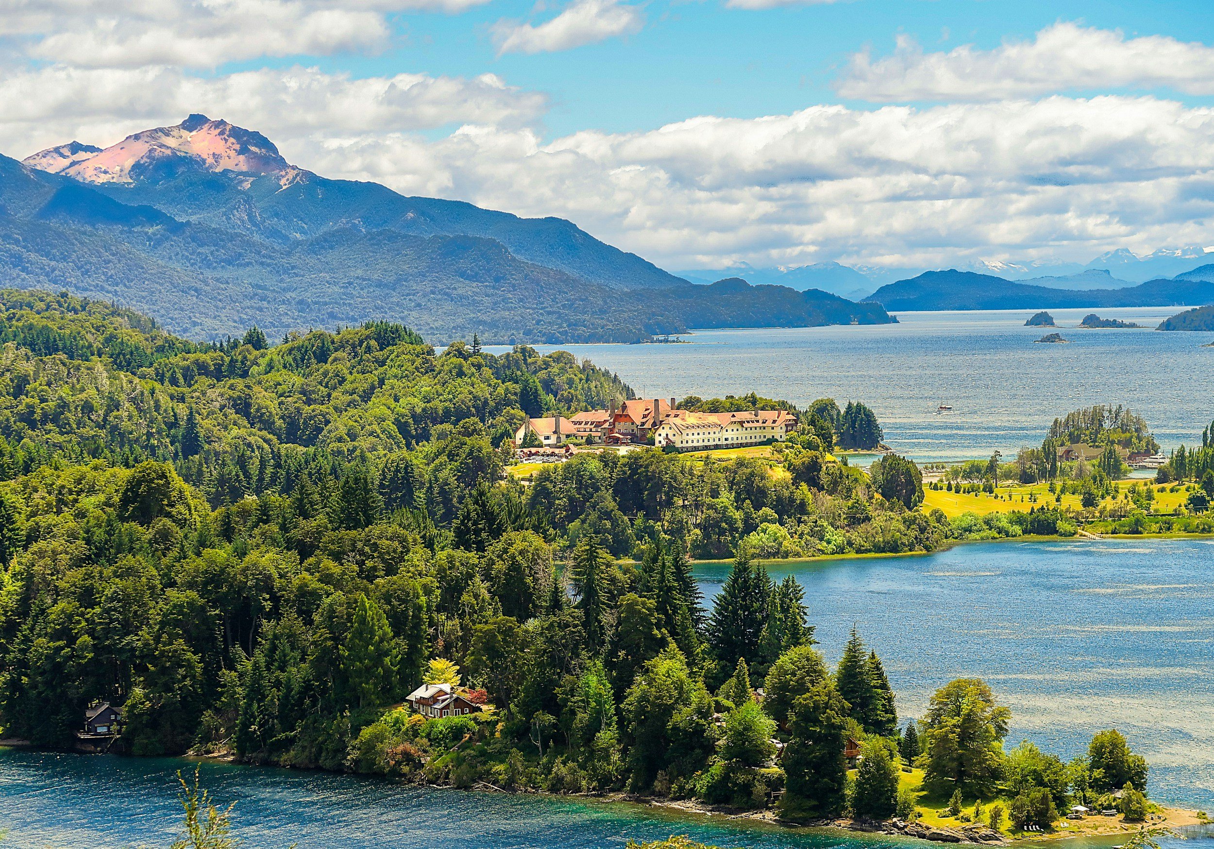 The turquoise Nahuel Huapi lake near Bariloche with a classic Swiss-style hotel on a wooded peninsula and snow-capped Andean peaks in the background, Argentina