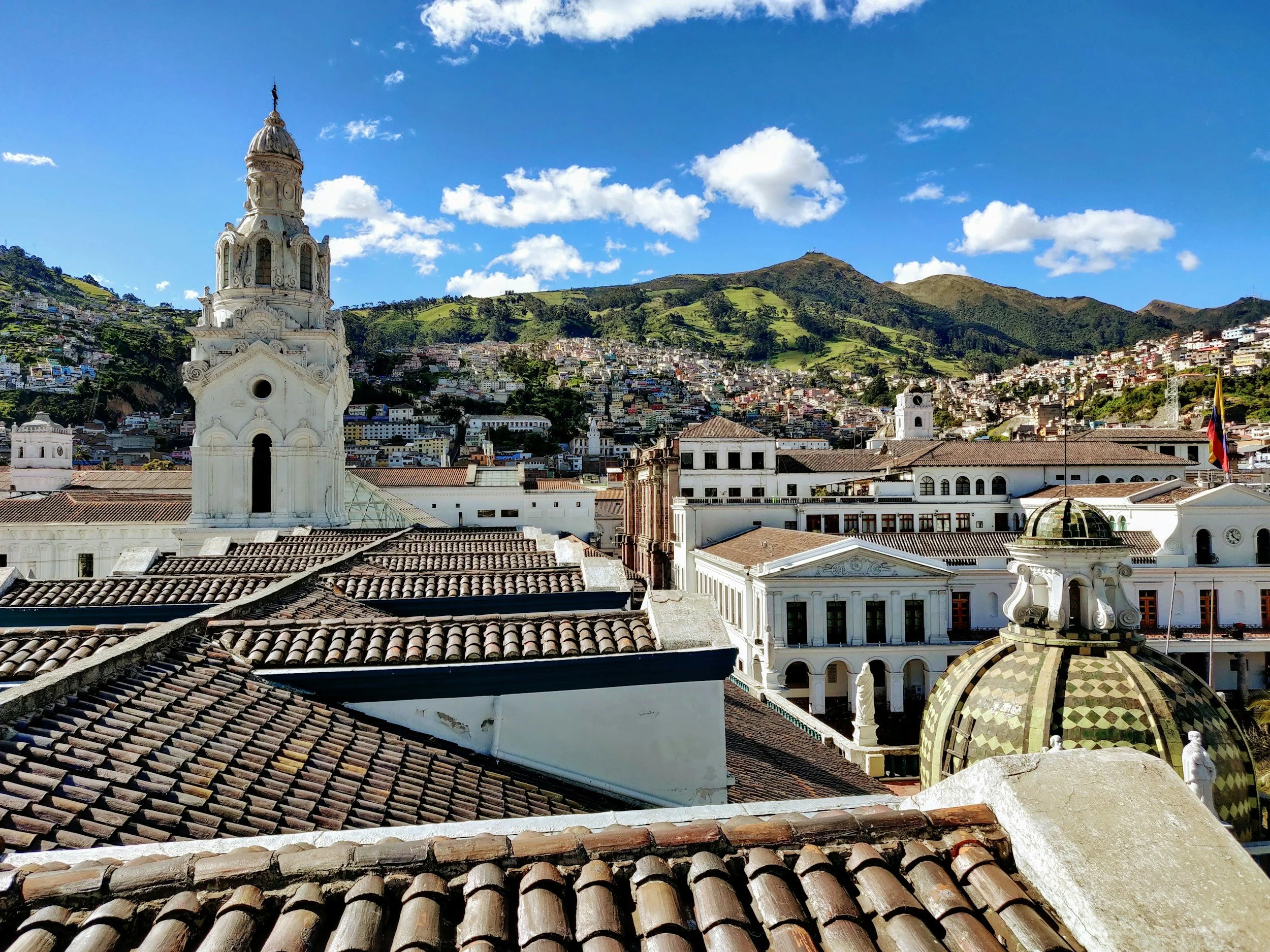 Aerial view over the colonial rooftops and baroque church towers of Quito historic centre with green Andean hills rising behind, Ecuador