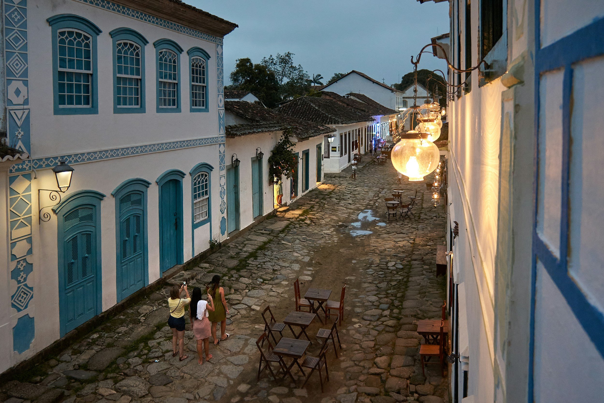 Travellers exploring a cobblestone street lined with blue-trimmed colonial buildings and hanging lanterns in Paraty, Brazil