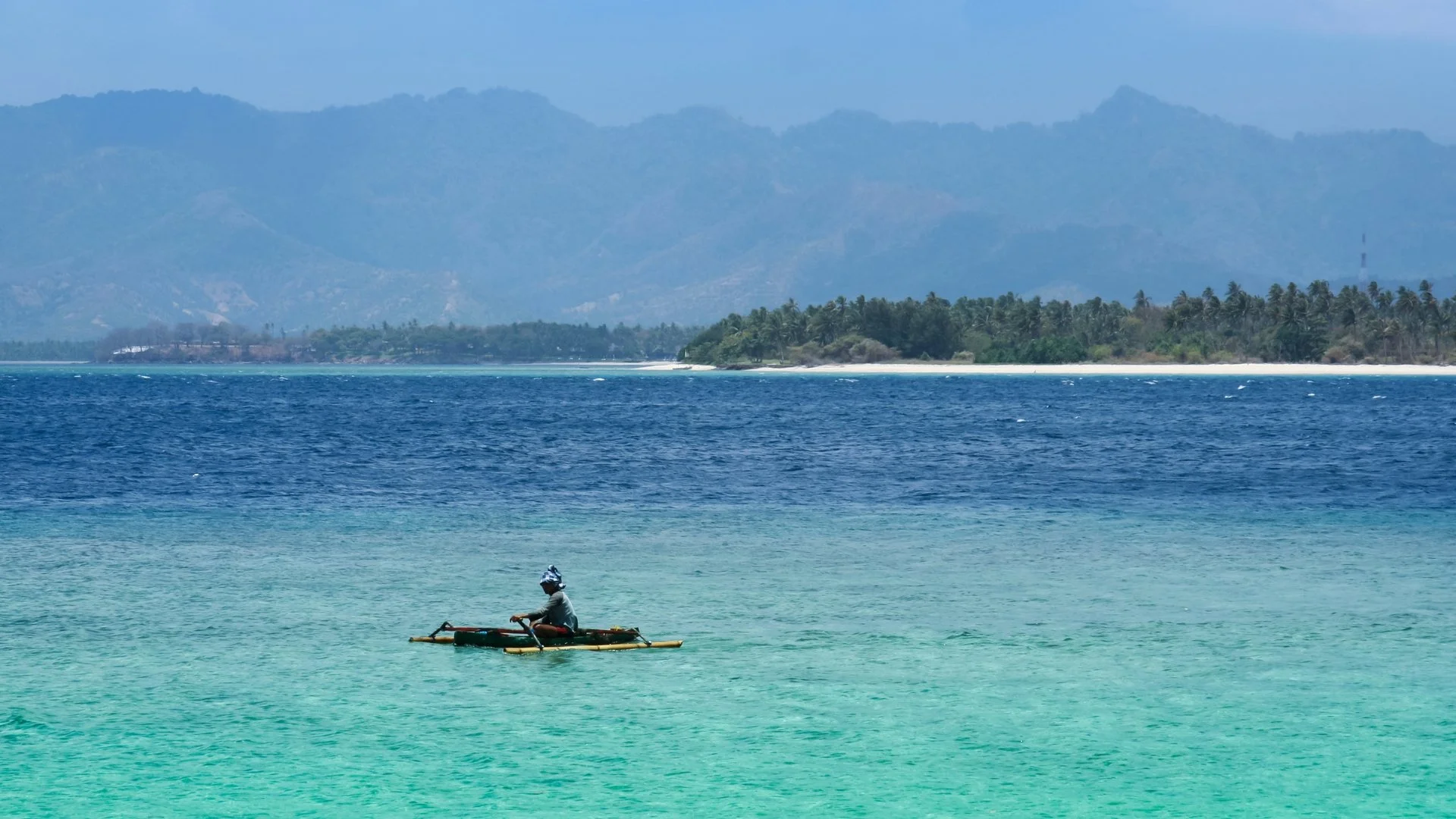 Local fisherman on a small traditional outrigger boat on calm turquoise waters off the coast of Lombok, Indonesia, with a forested island visible in the distance