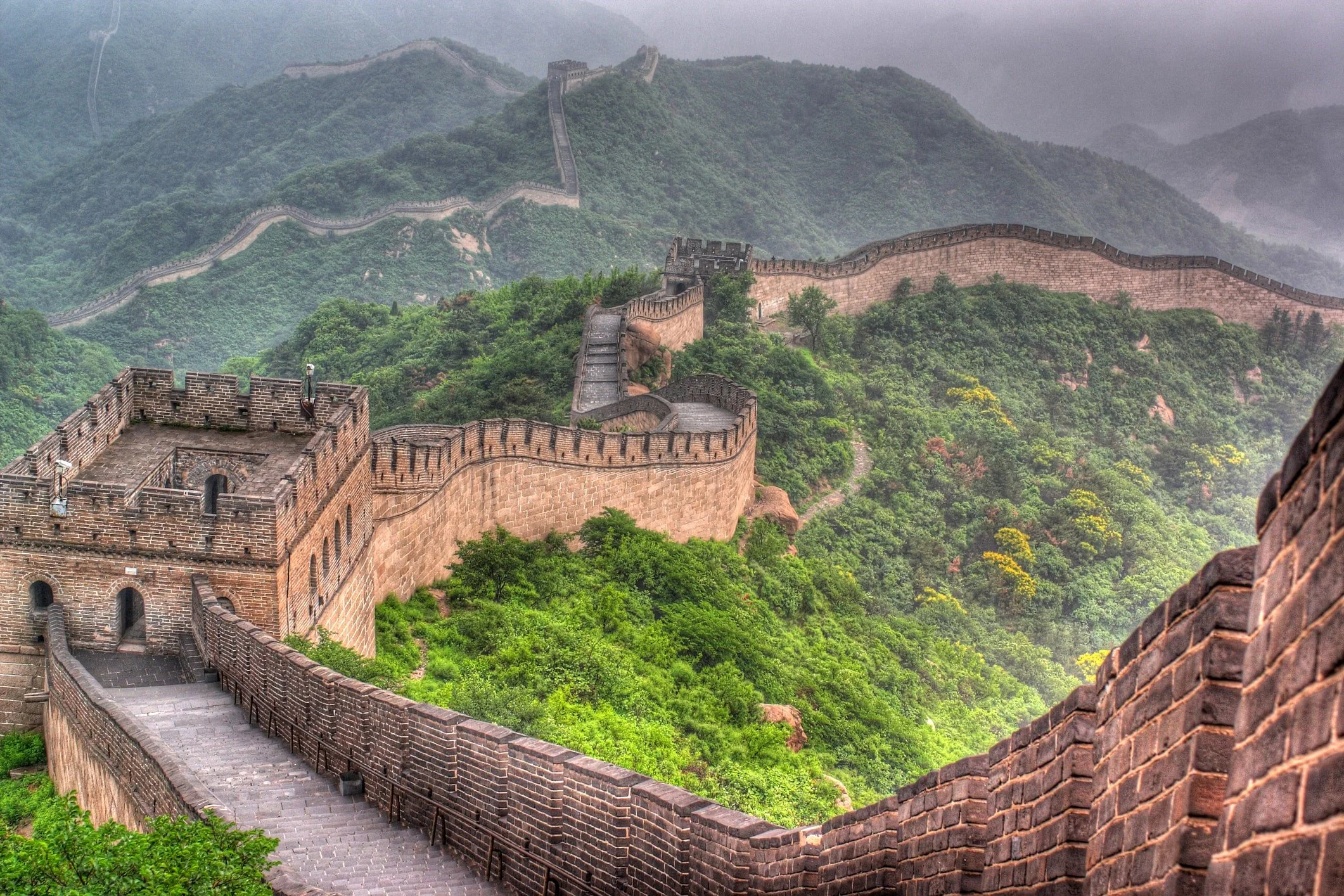 The Great Wall of China winding through densely forested green hills in summer, with a watchtower in the foreground and the wall stretching into the misty distance