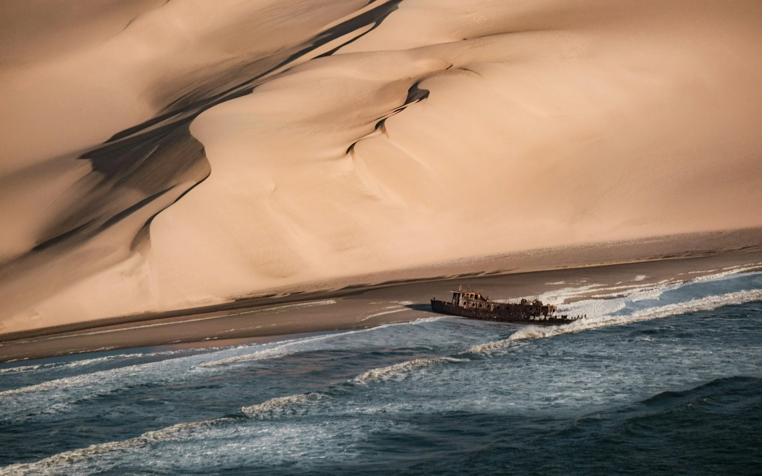 A rusting shipwreck stranded on the shore of the Skeleton Coast in Namibia, with sweeping desert sand dunes curving to meet the Atlantic Ocean surf in the background