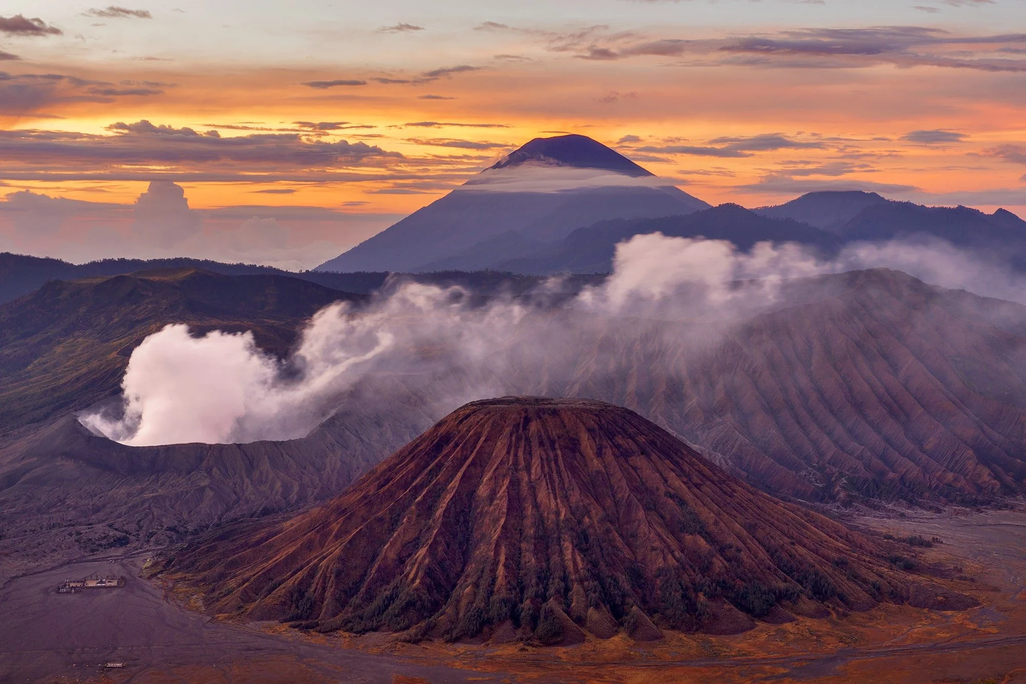 Mount Bromo active volcano rising from a sea of cloud at sunrise with the distant peak of Mount Semeru silhouetted against an orange and purple sky in East Java, Indonesia
