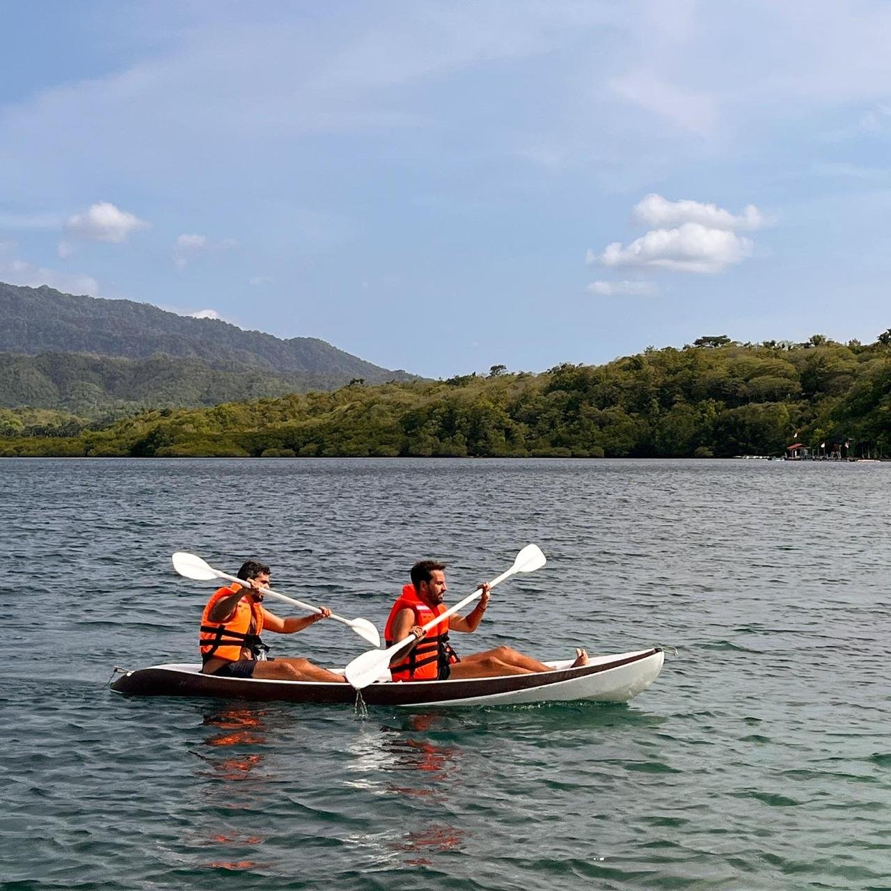 Two people kayaking on a body of water with a forested shoreline and hills in the background, under a partly cloudy sky.