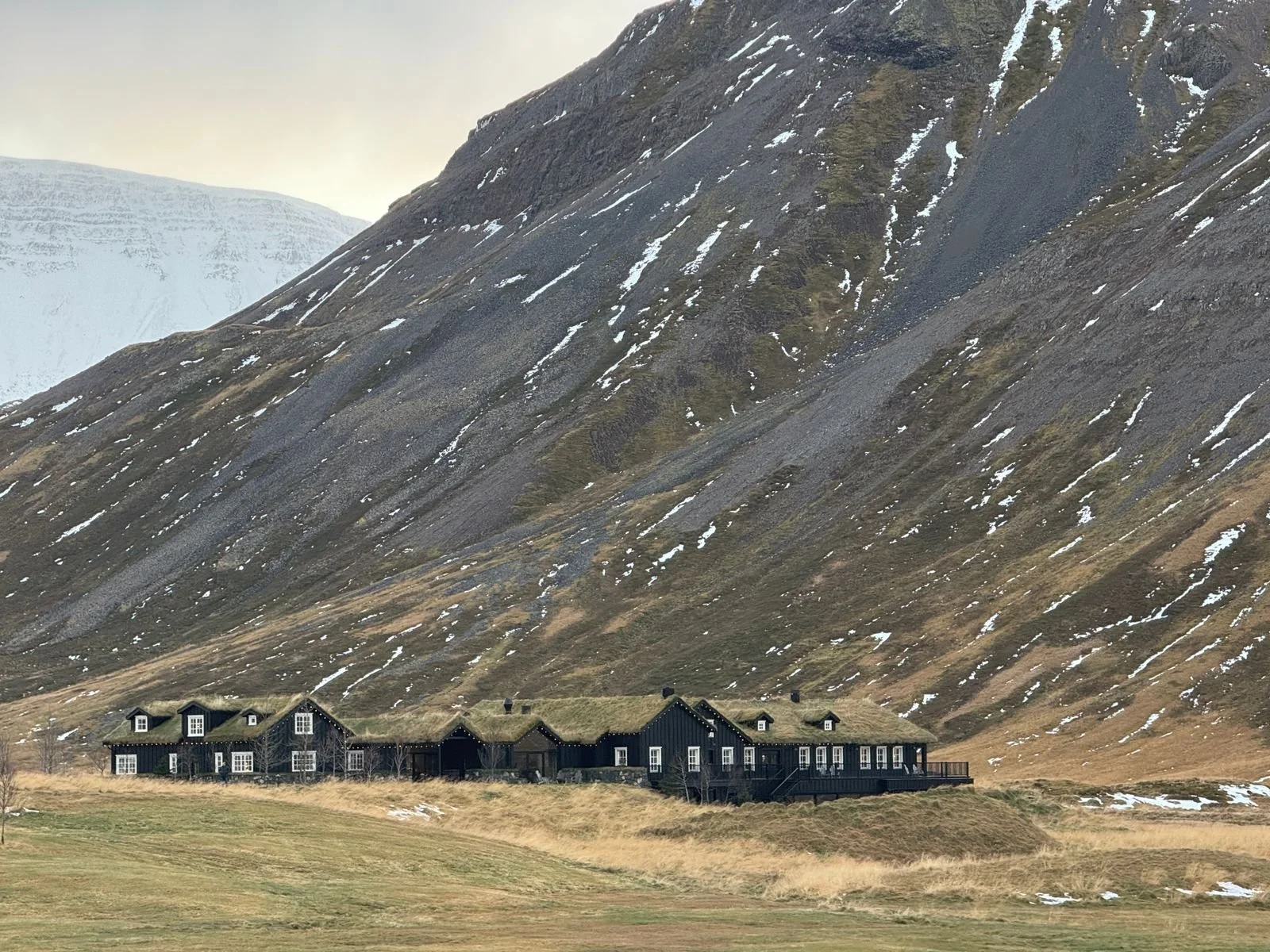 Deplar Farm luxury lodge nestled below a vast snow-streaked mountain in the remote Fljót valley of northern Iceland, with dark timber buildings set against the dramatic landscape
