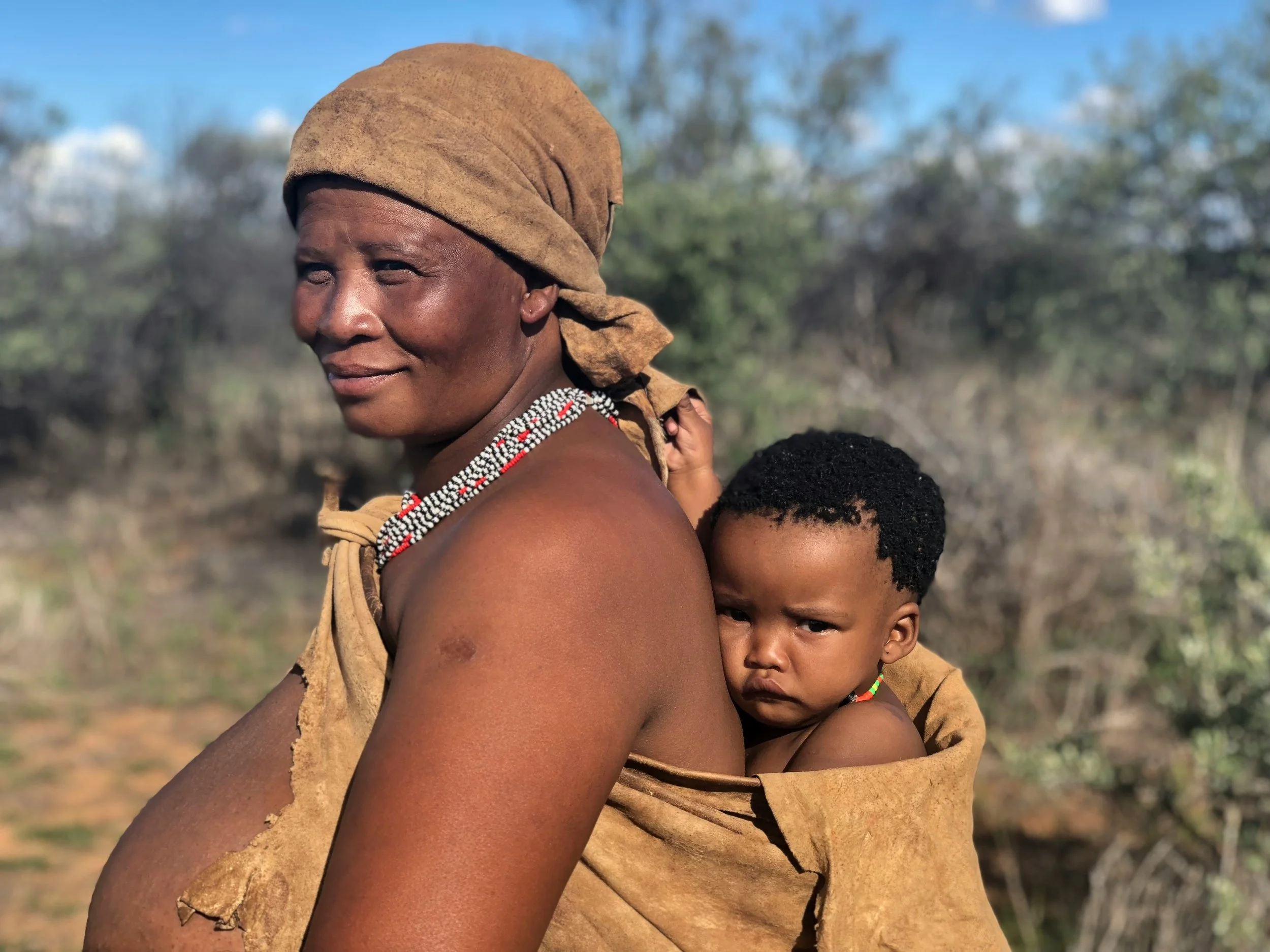 A Himba woman wearing ochre-covered traditional dress and head wrap carrying a young child on her back in rural Namibia, with dry acacia scrubland visible in the background