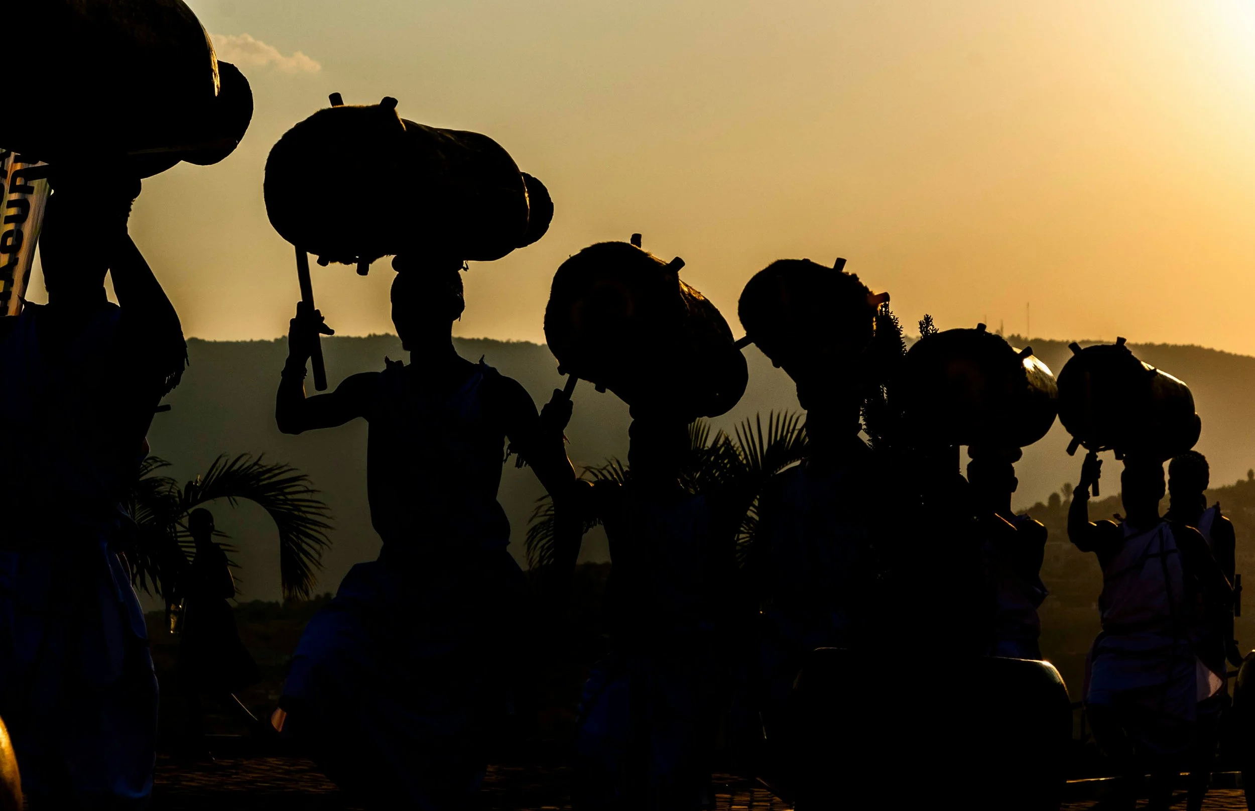 Silhouettes of traditional Intore drummers holding large drums above their heads against a golden sunset sky in Rwanda, with palm trees and rolling hills in the background