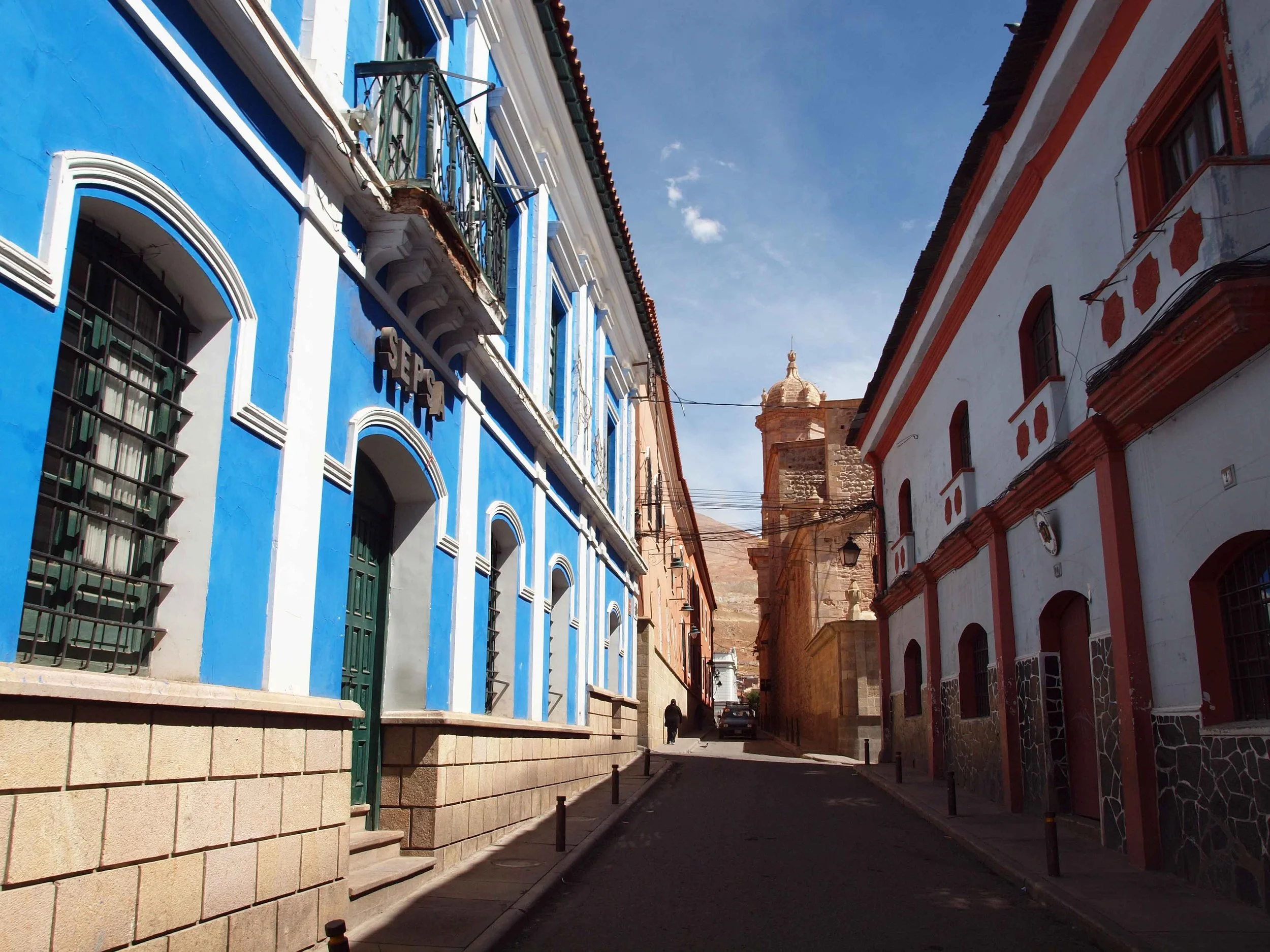 Narrow colonial street in Potosí lined with blue and white buildings and a baroque church tower in the background, Bolivia