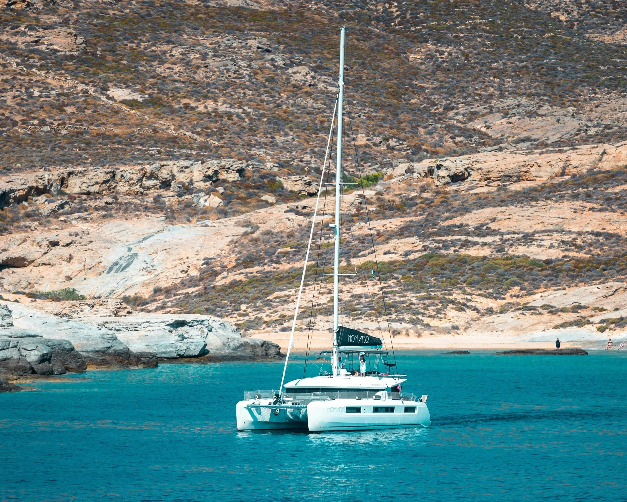 A private catamaran yacht sailing through turquoise water past a rocky uninhabited Cycladic island, on a private sailing tour through the lesser-known islands of Greece