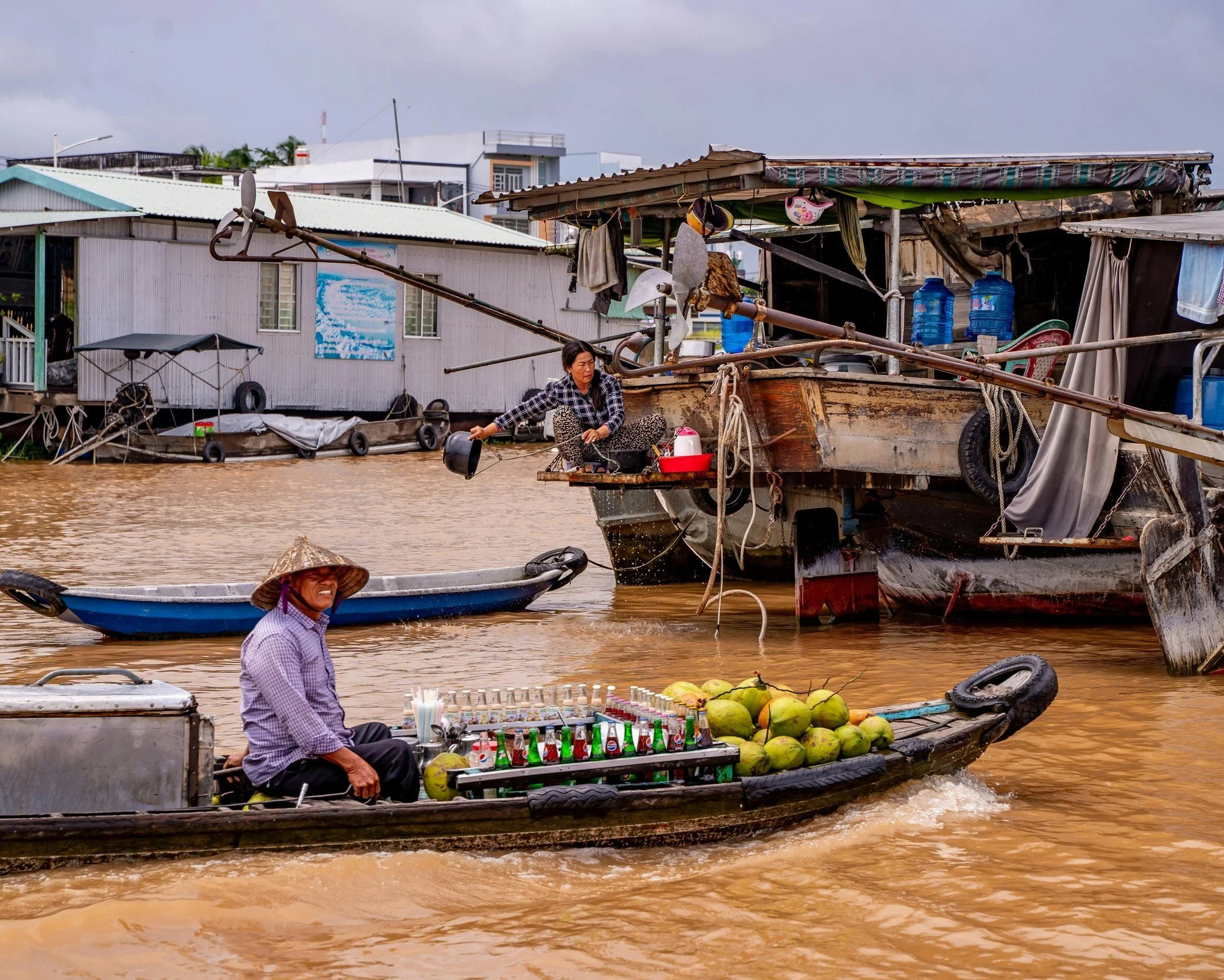 A woman in a conical hat sitting on a wooden boat loaded with coconuts and produce at a floating market in the Mekong Delta, Vietnam, with houseboats and water vendors visible behind her